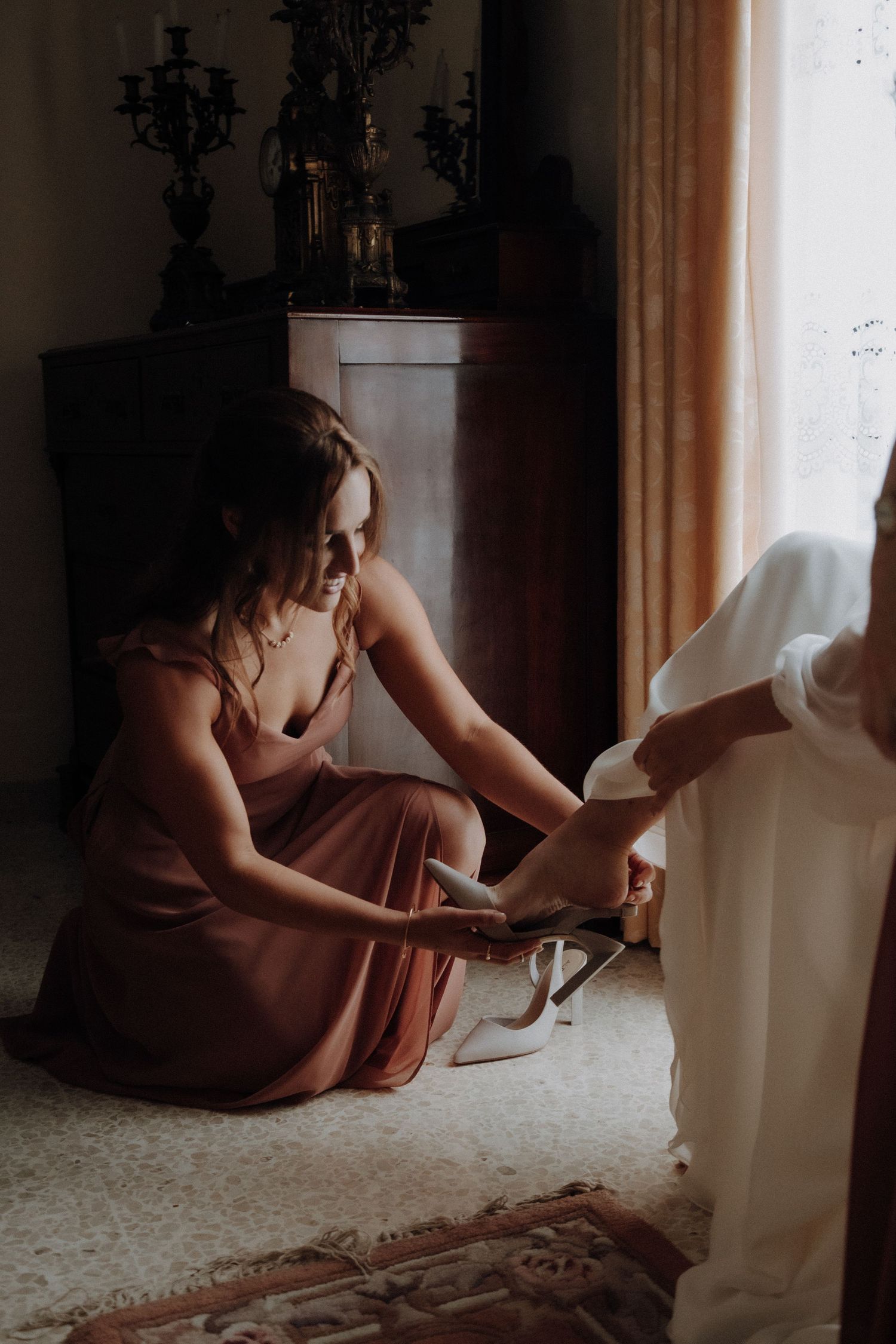 Bridesmaid in burgundy dress helping with bridal shoes in vintage room setting.