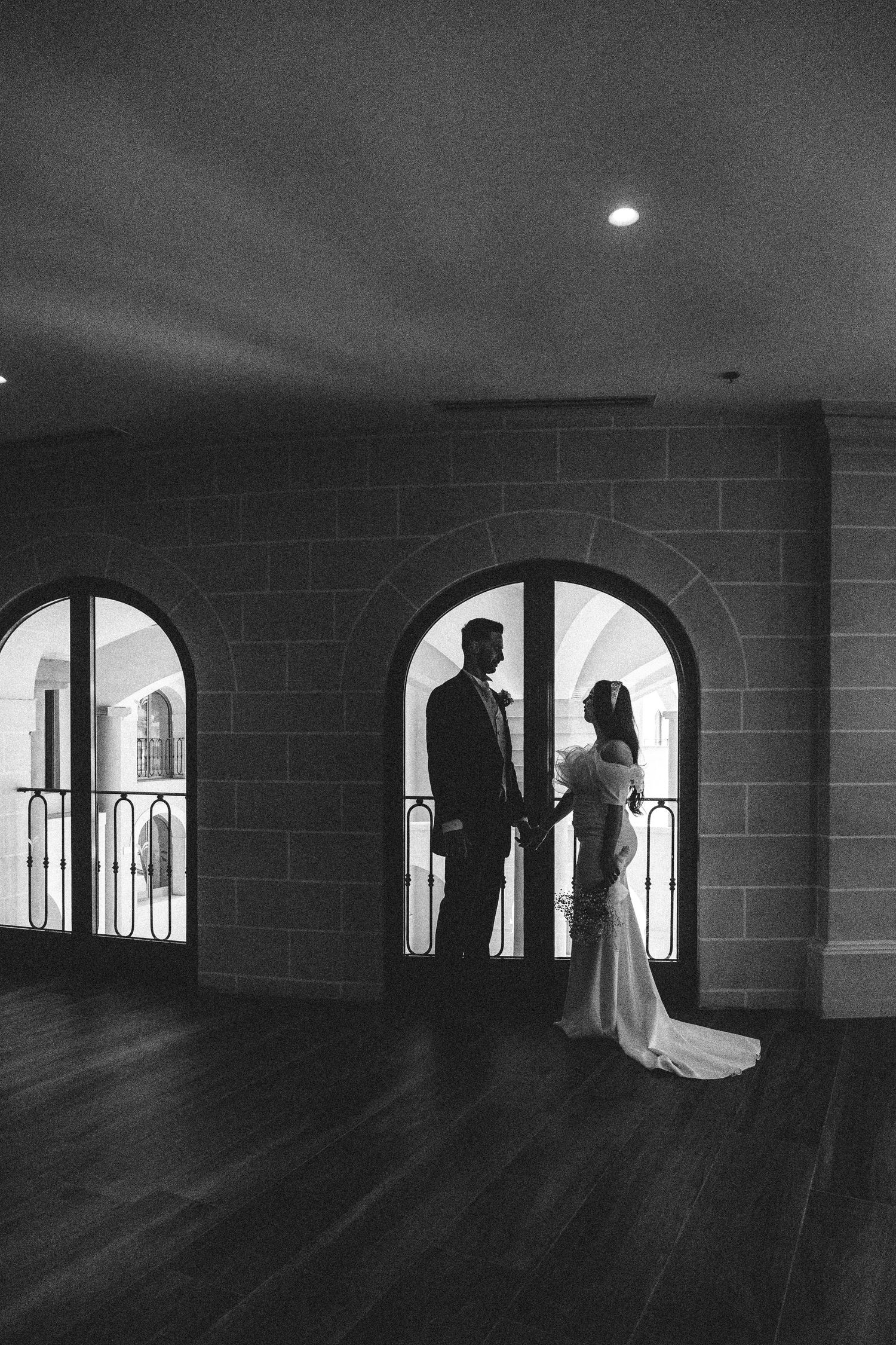 Newlyweds share intimate moment in arched window alcove overlooking cityscape.