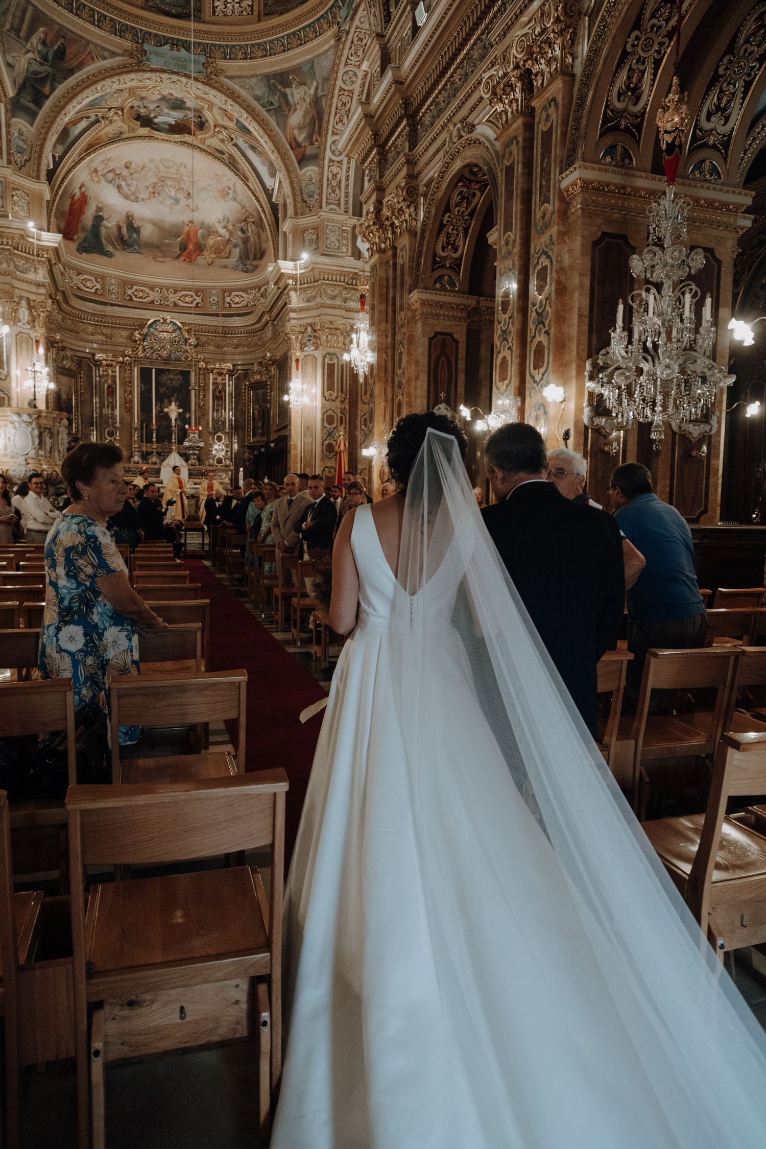 Bride in flowing white gown and veil walks down ornate church aisle with baroque ceiling.