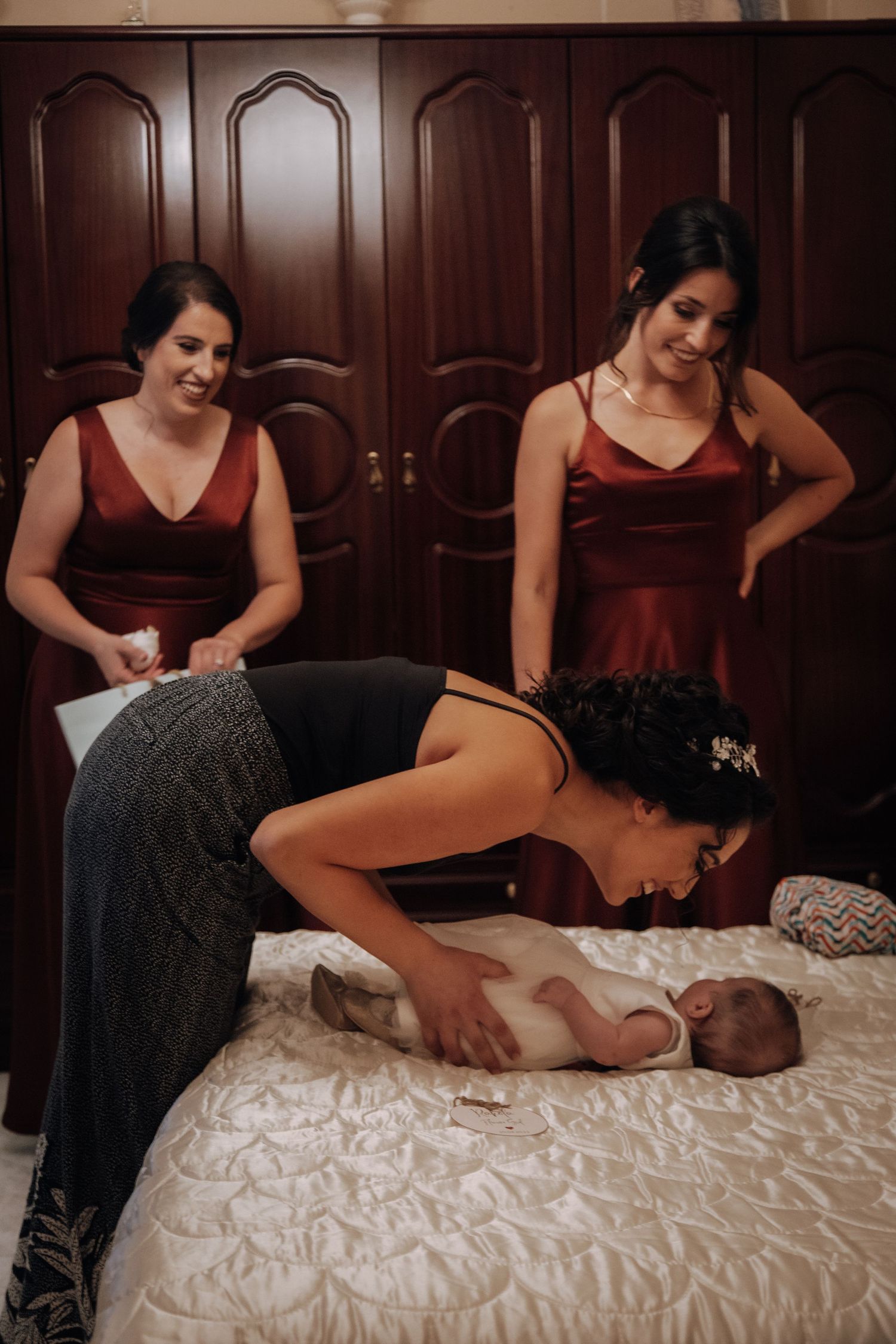 A baby lies on a white quilted bed while someone leans over them, with two figures in burgundy dresses watching.