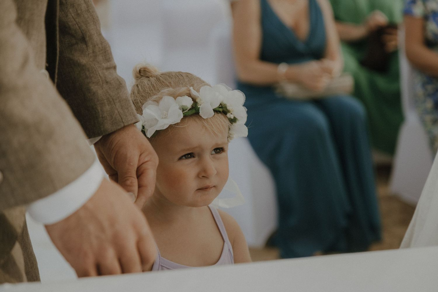 Young flower girl in white dress with floral crown watches wedding ceremony.