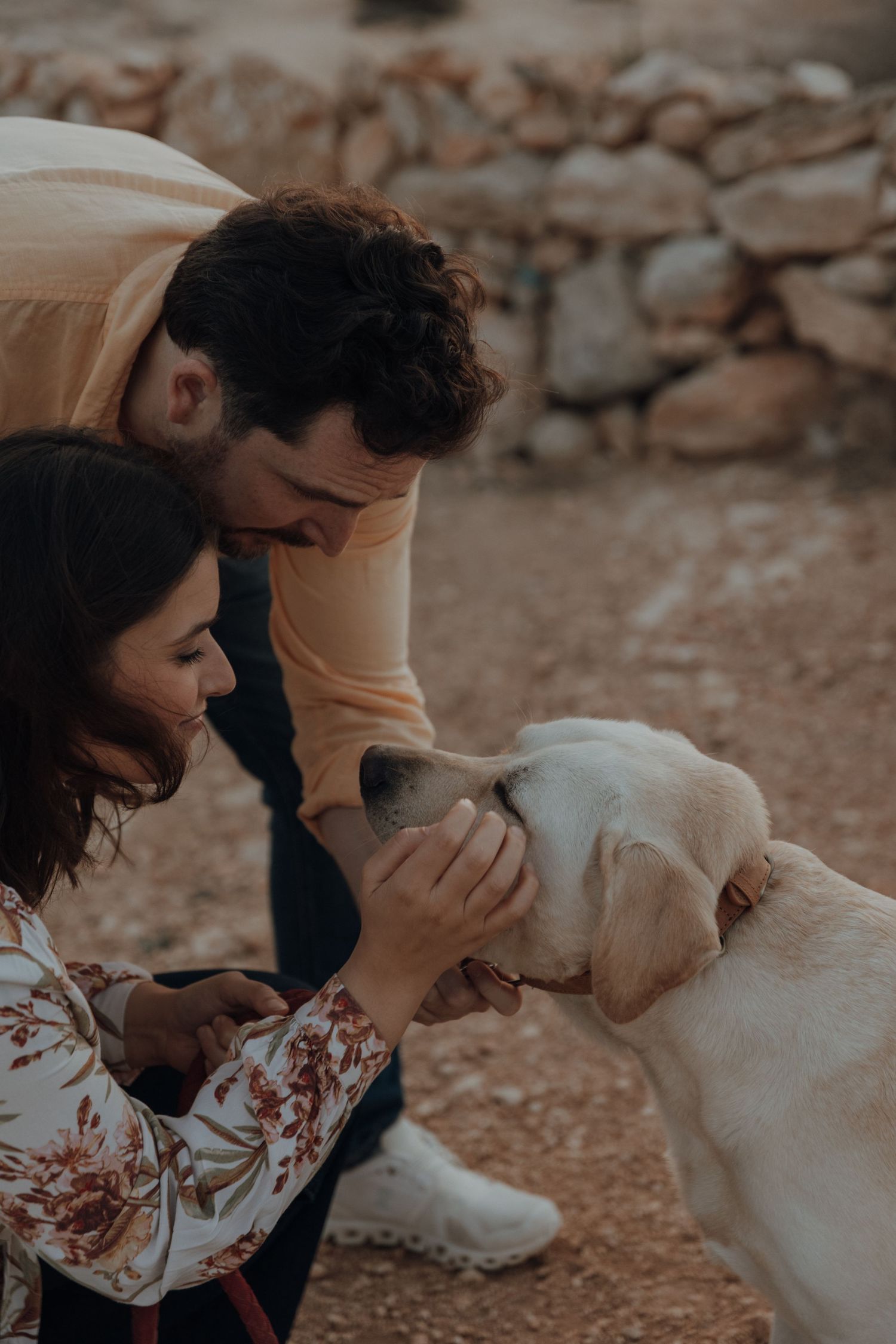 A couple plays with their white dog on rocky terrain.