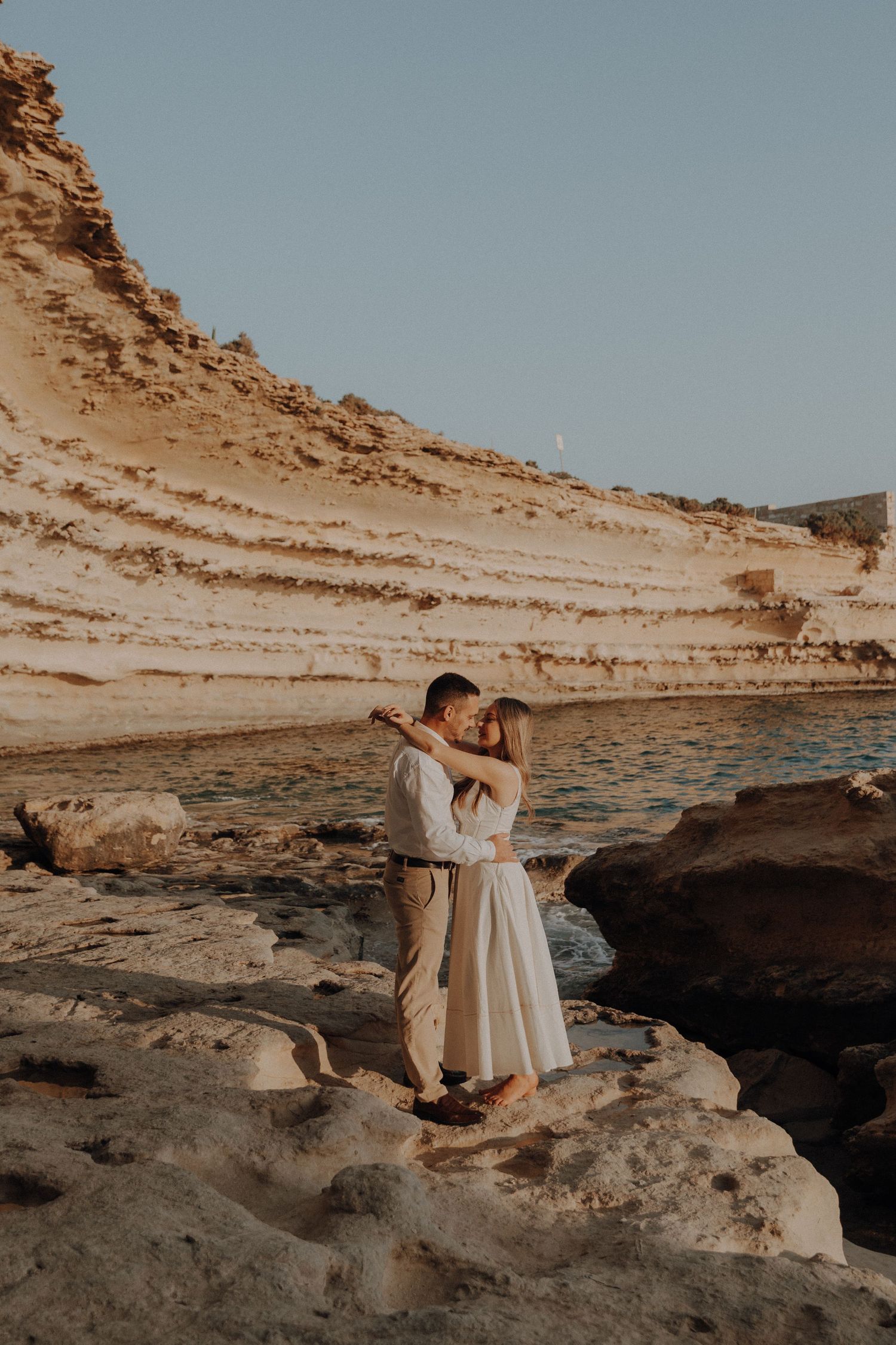 Couple embracing romantically on rocky shore with layered sandstone cliffs and calm water in warm golden light.