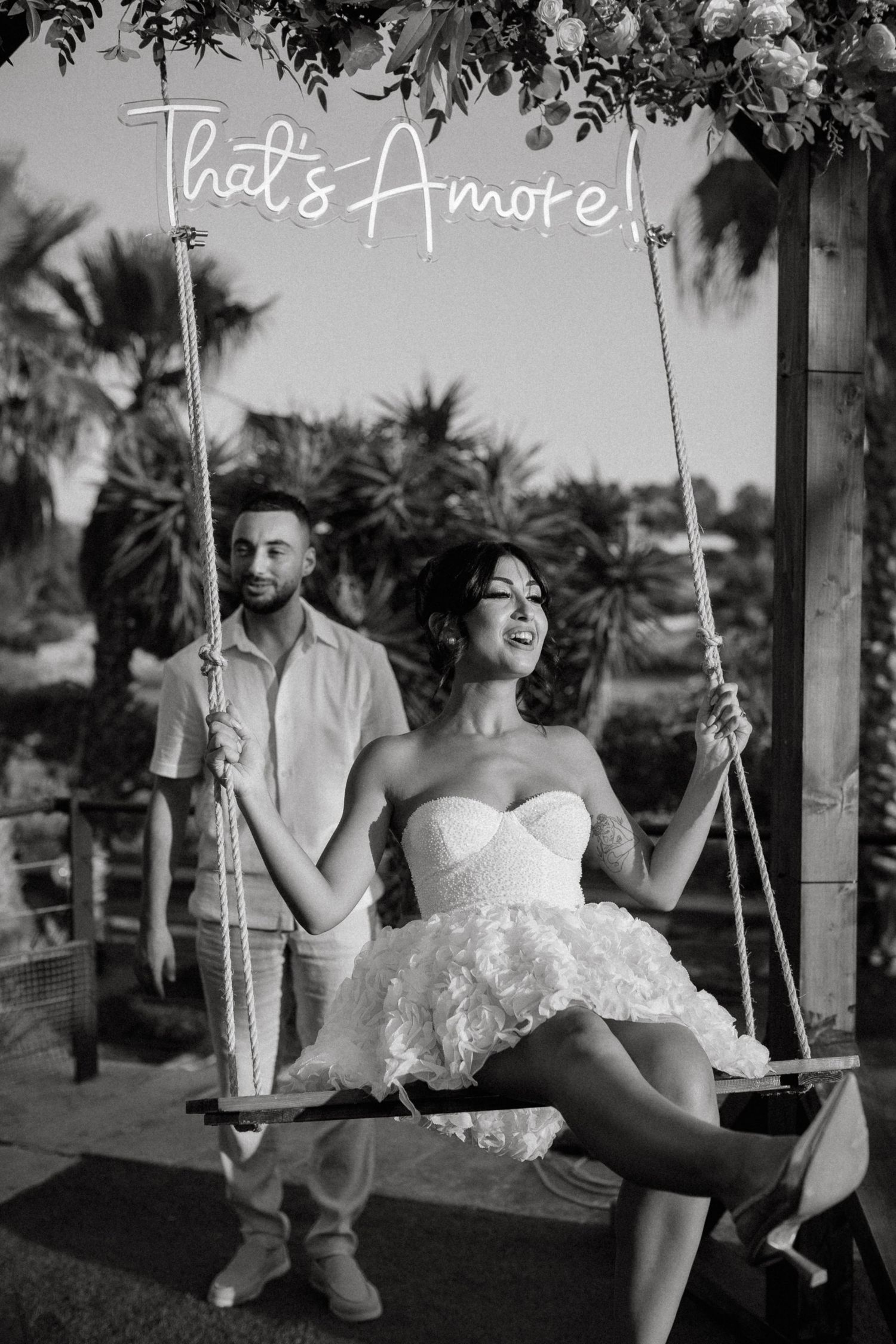 A romantic black and white photo of a couple by a neon 'That's Amore' sign with palm trees in the background.