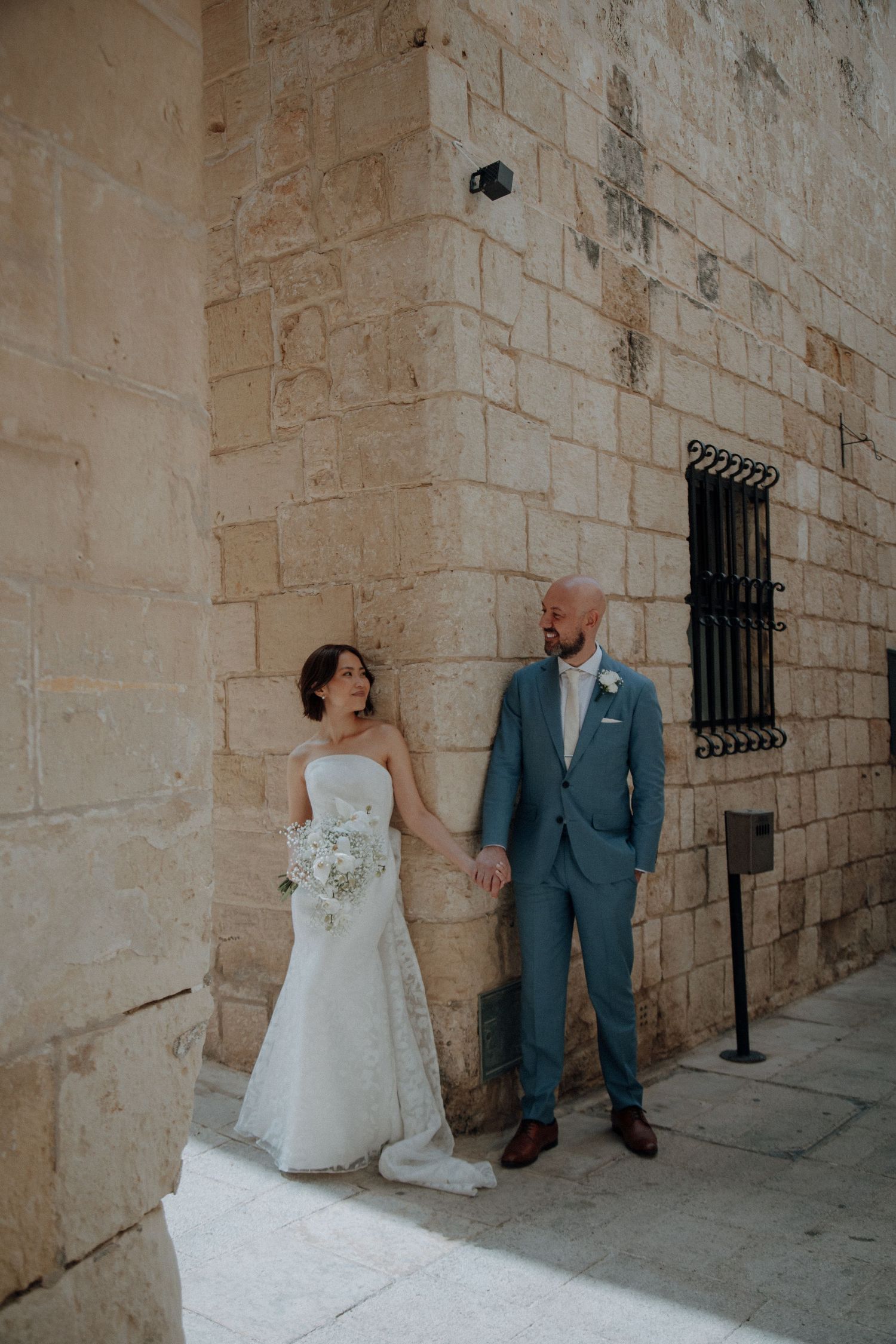 A full-length wedding portrait against a stone wall showcasing a white gown and blue suit.