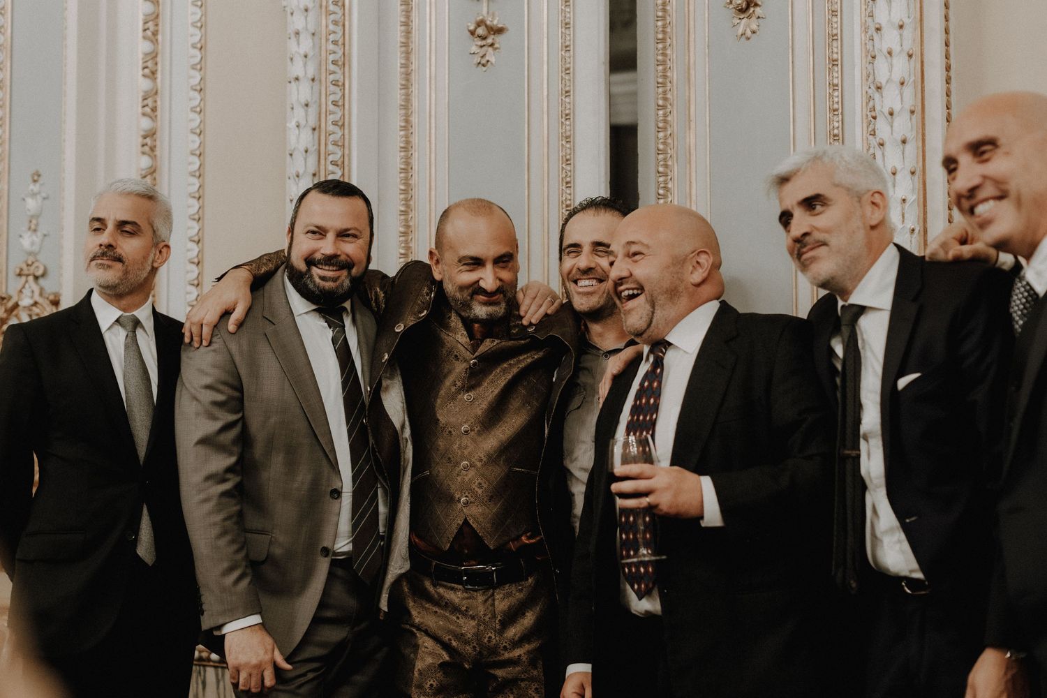 Group of men in suits posing together at a formal event against an ornate wall.