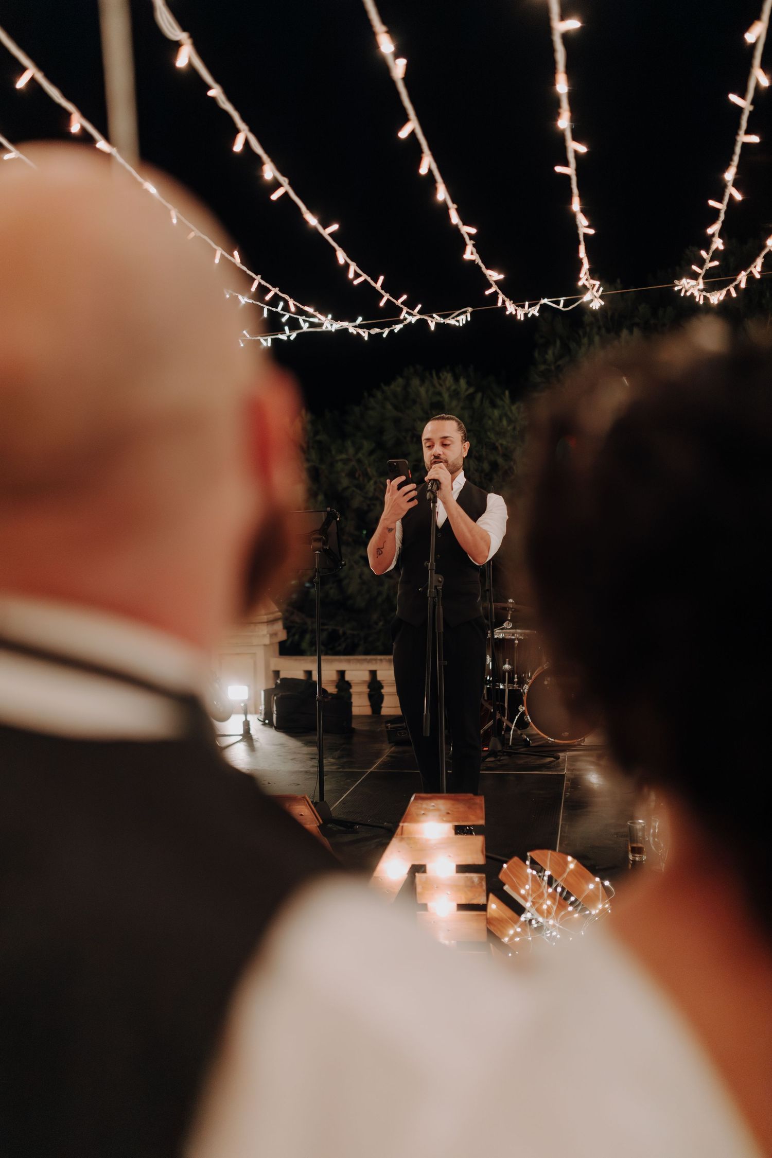 Live performer sings on stage beneath string lights at outdoor wedding reception.