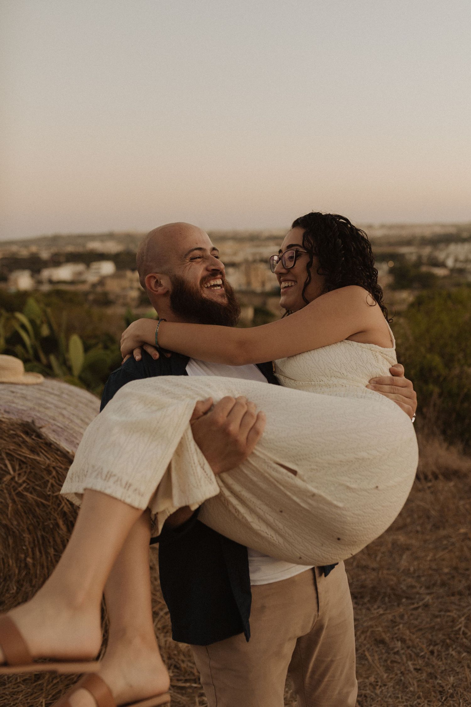 A couple shares a lighthearted moment against the backdrop of a Mediterranean sunset landscape.