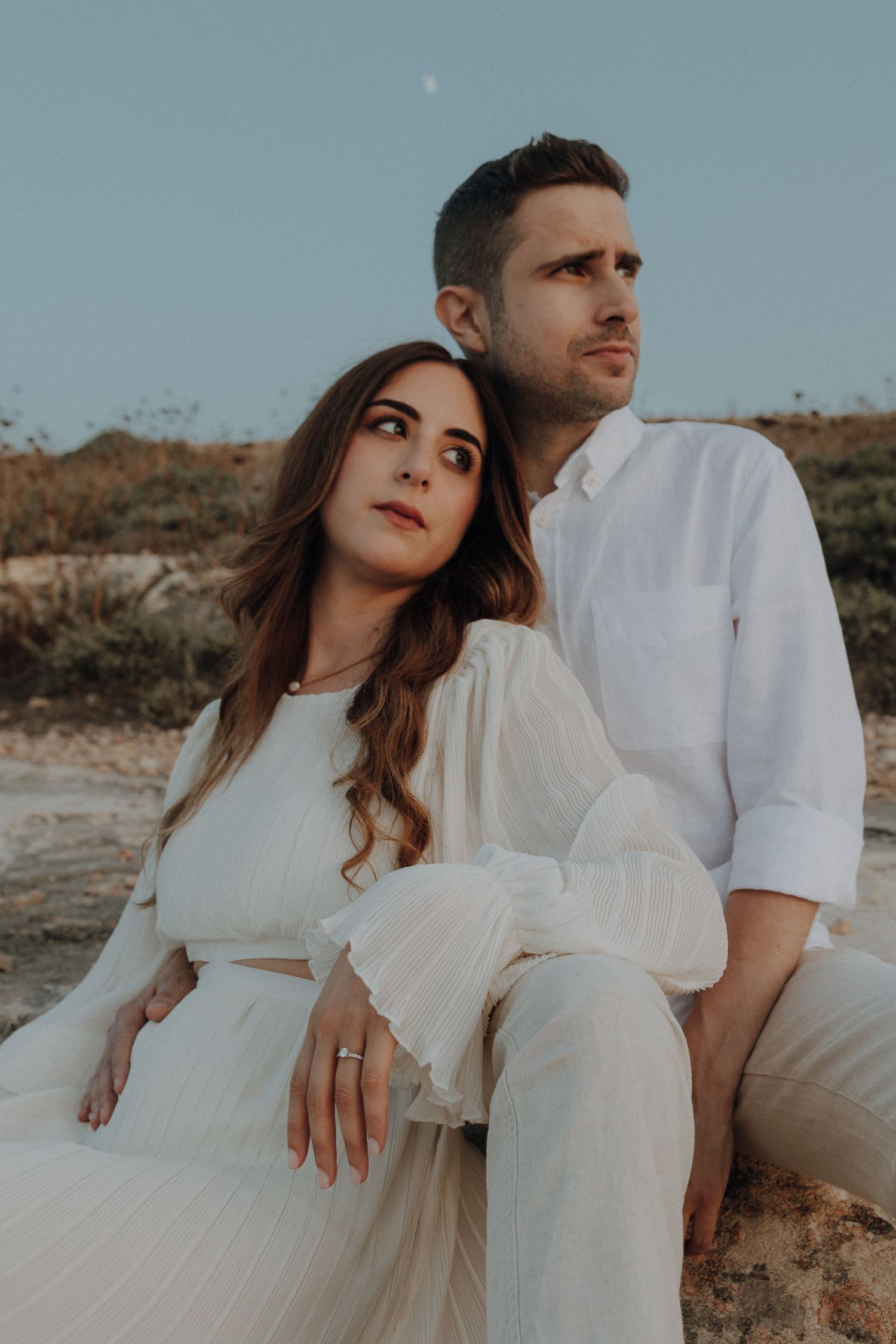 Romantic engagement photo session with couple in white outfits on desert landscape.