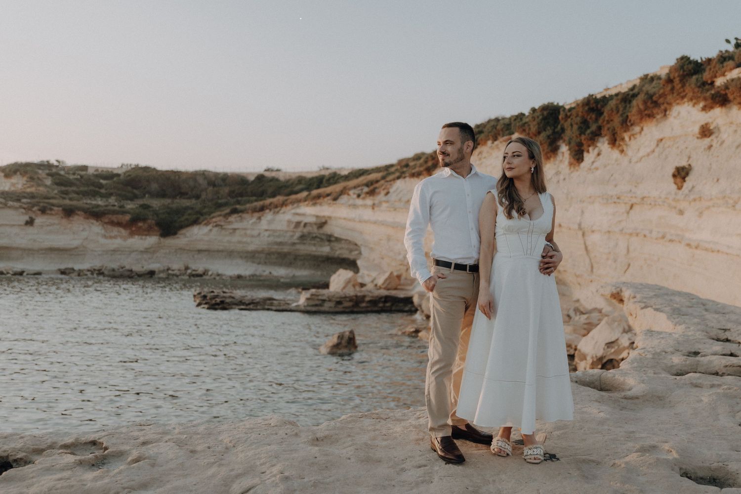 Happy couple walking along rocky coastal cliffs at golden hour, woman in white dress, man in casual attire.