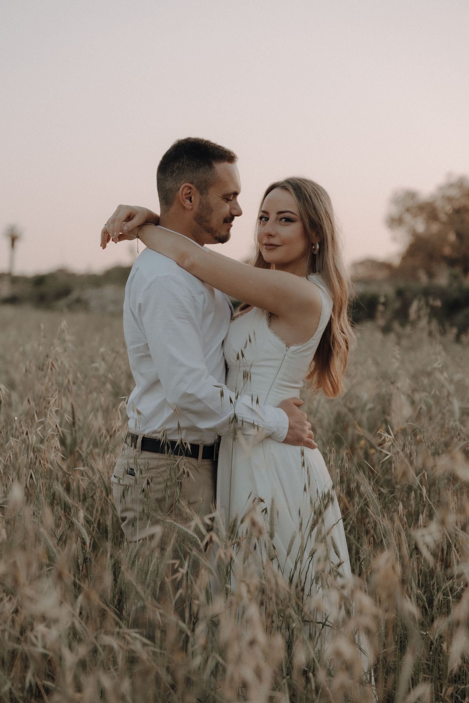 Couple embracing romantically in a golden wheat field at sunset, woman in white dress with arms around mans neck.