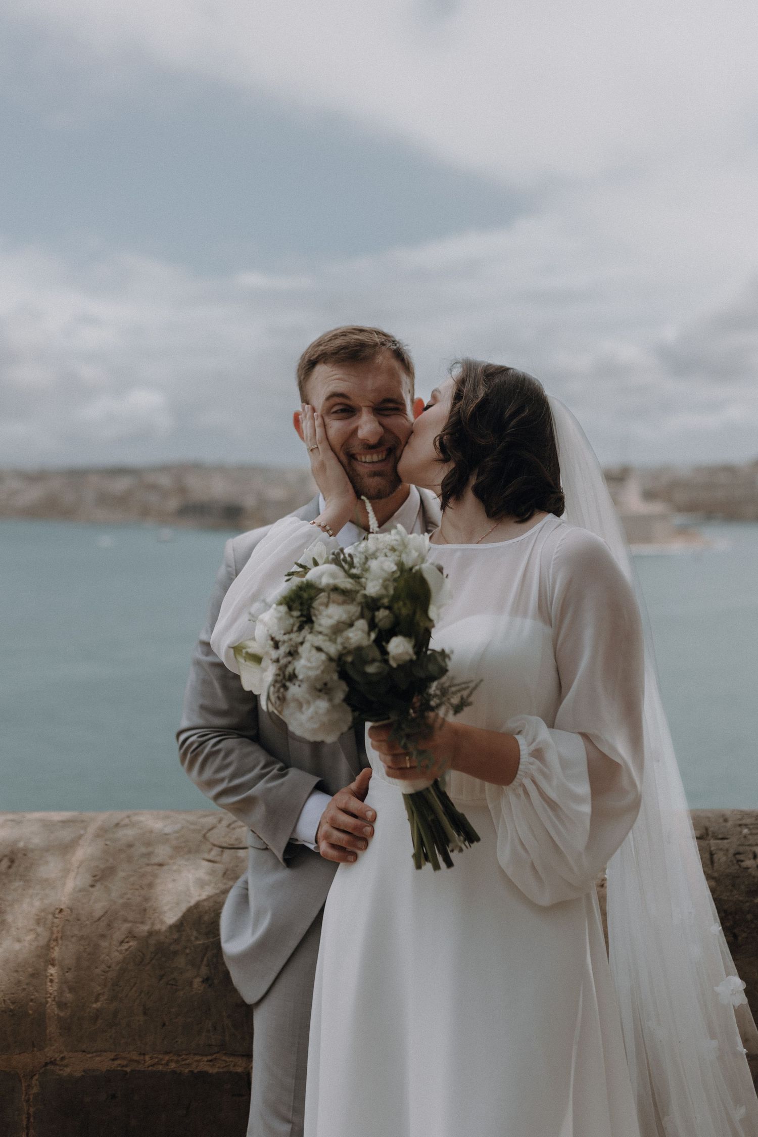 A couple shares an intimate moment by the ocean while holding a white floral bouquet.