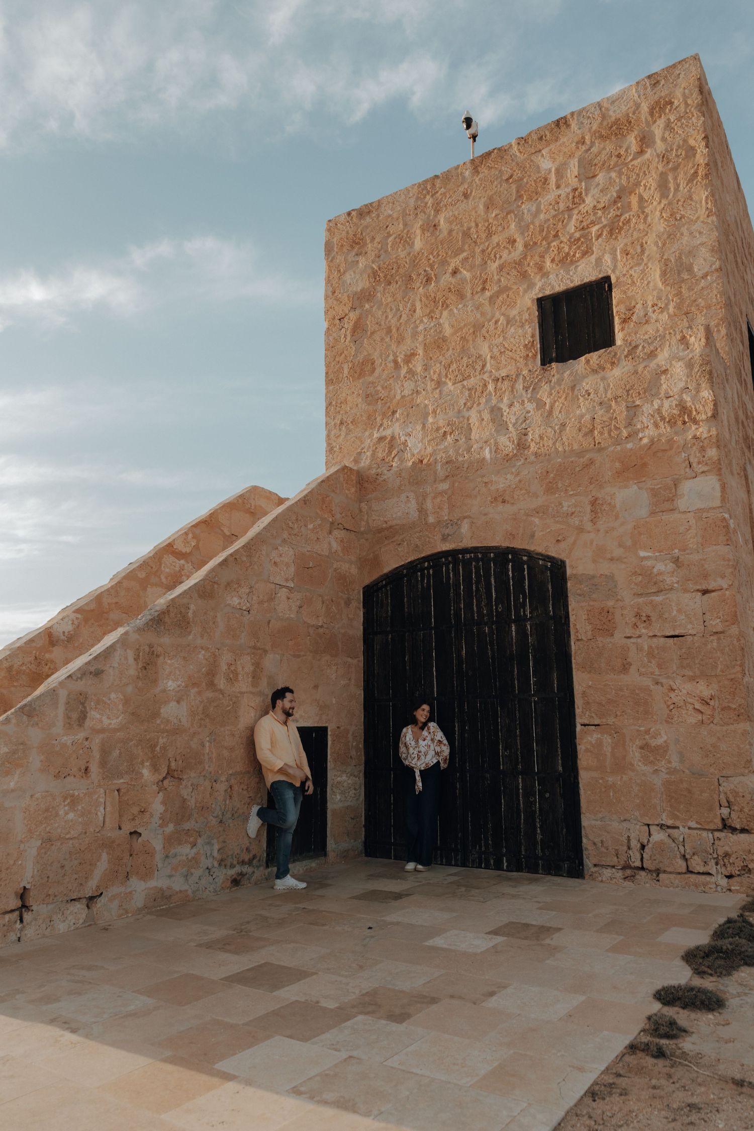 Two people stand outside a rustic stone fortress entrance with large black wooden doors under cloudy skies.