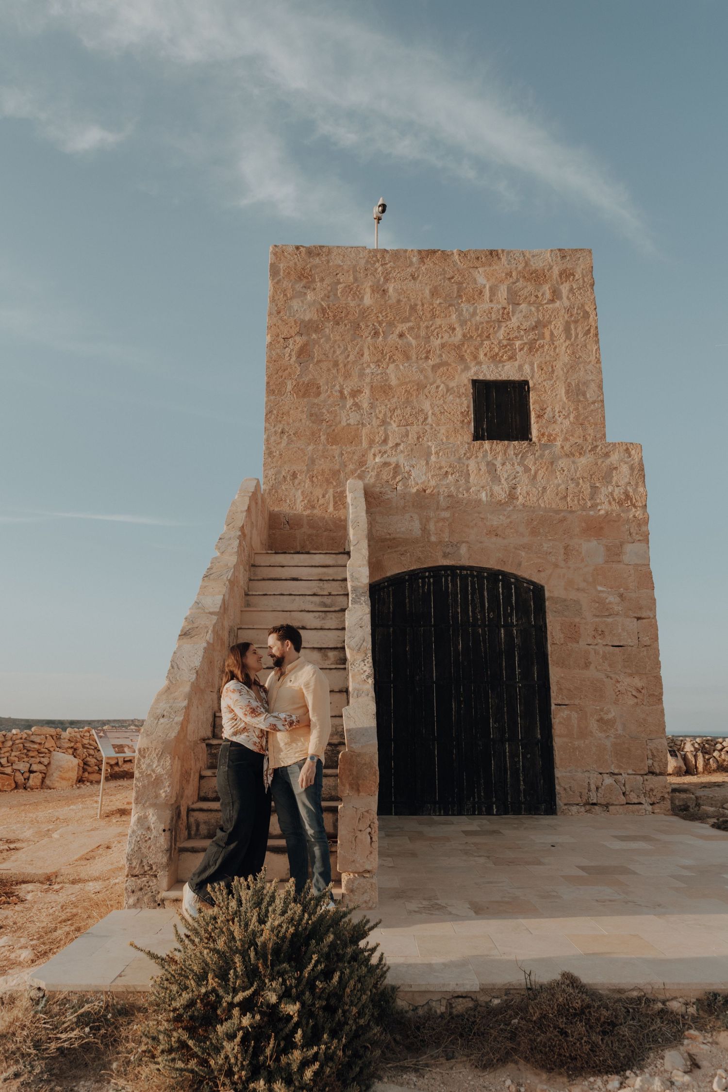 A romantic couple embraces against a rustic stone building with an arched doorway in a desert landscape.