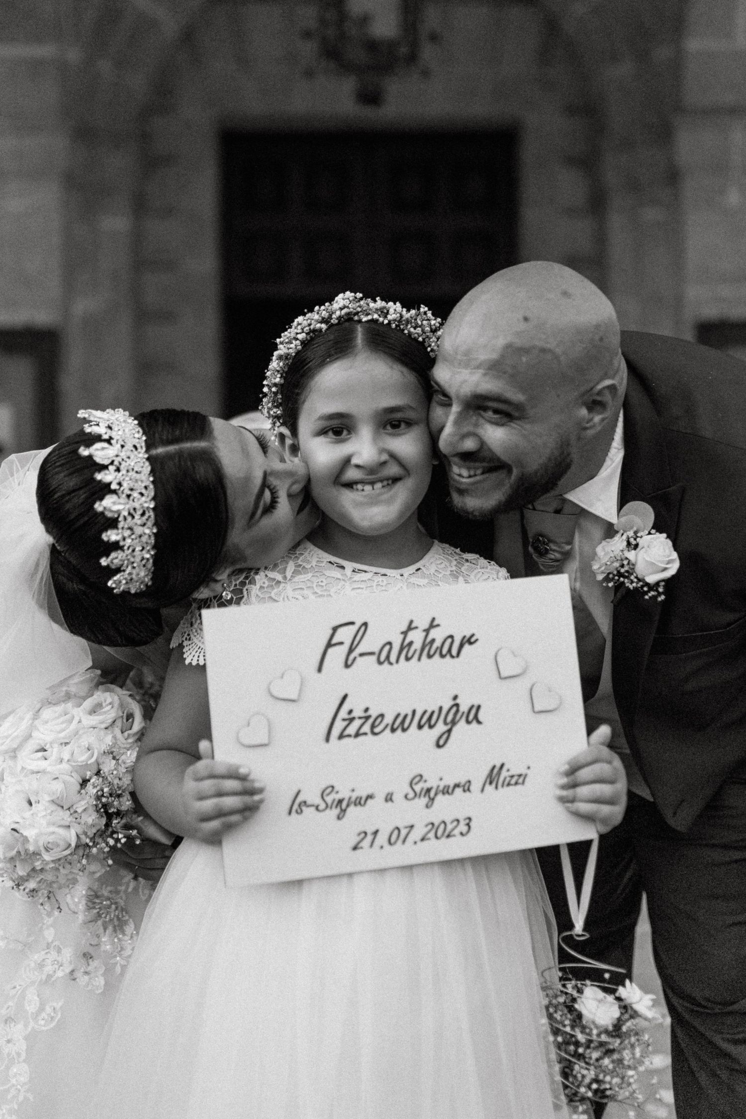 A young flower girl holds a sign with wedding details as couple kisses her cheeks.