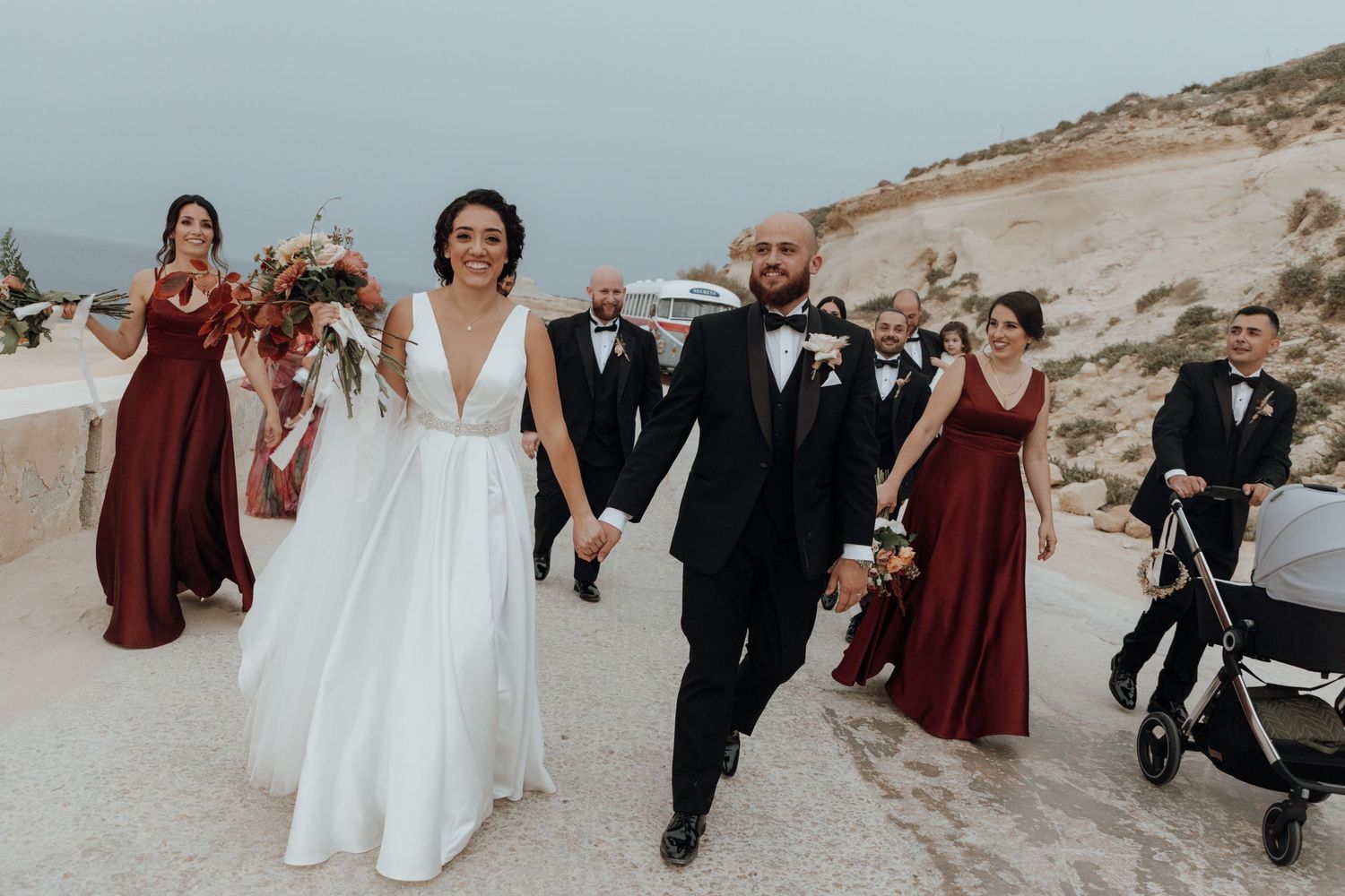 Wedding party walks along scenic coastal path wearing burgundy bridesmaid dresses and formal attire.