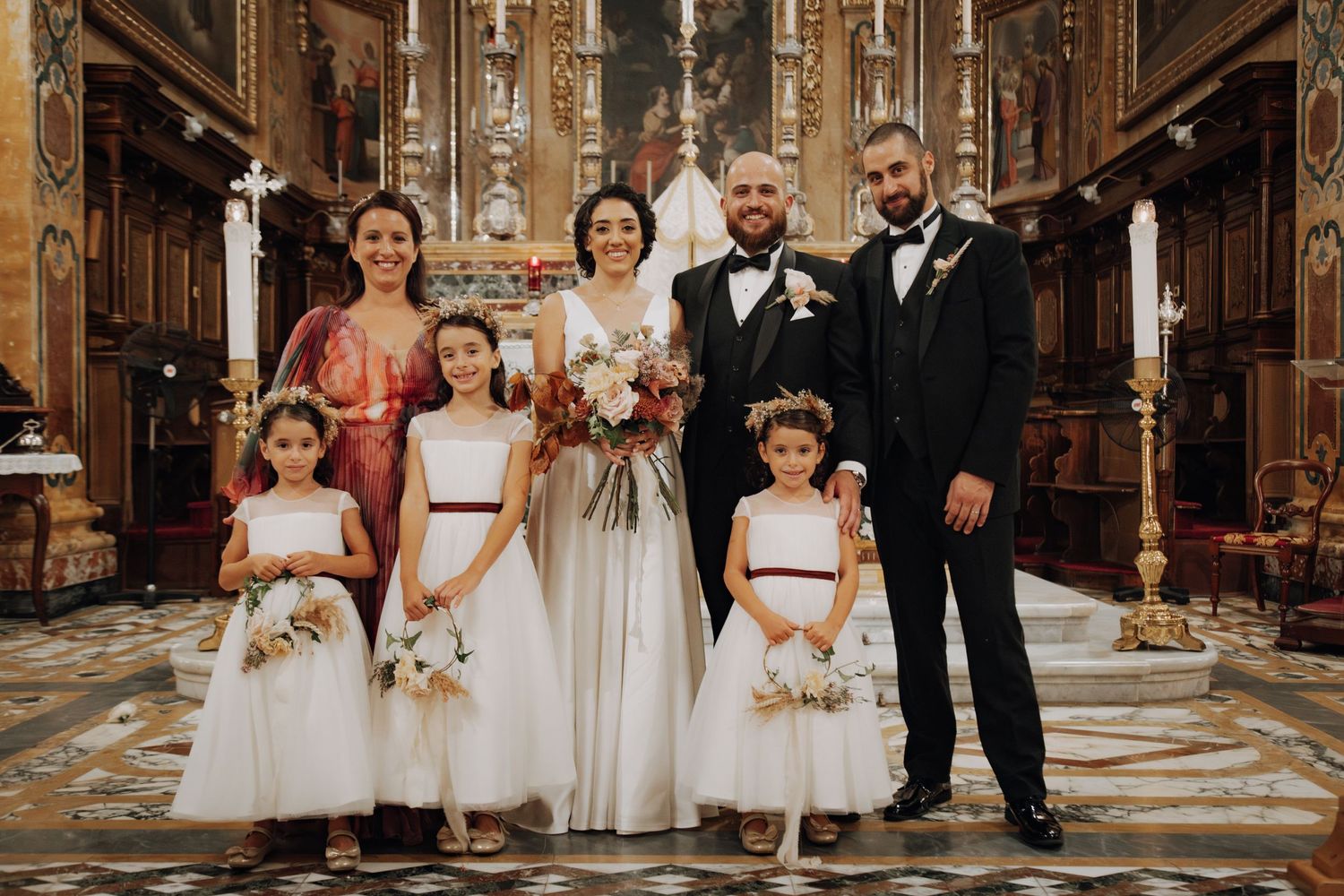 Bridal party with flower girls in white dresses gather for formal photo in ornate church setting.