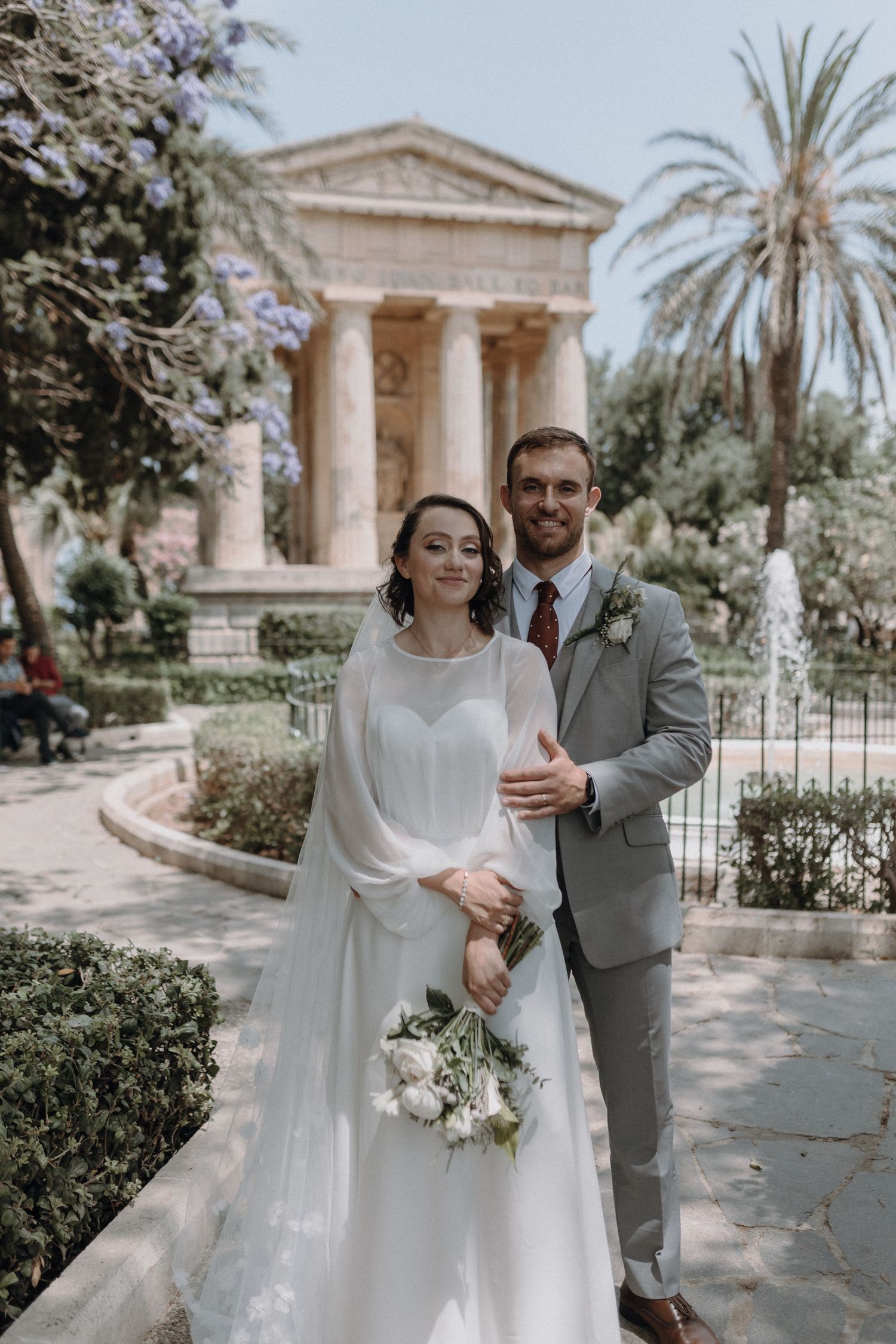 Newlyweds stand in front of a neoclassical building surrounded by palm trees and gardens.
