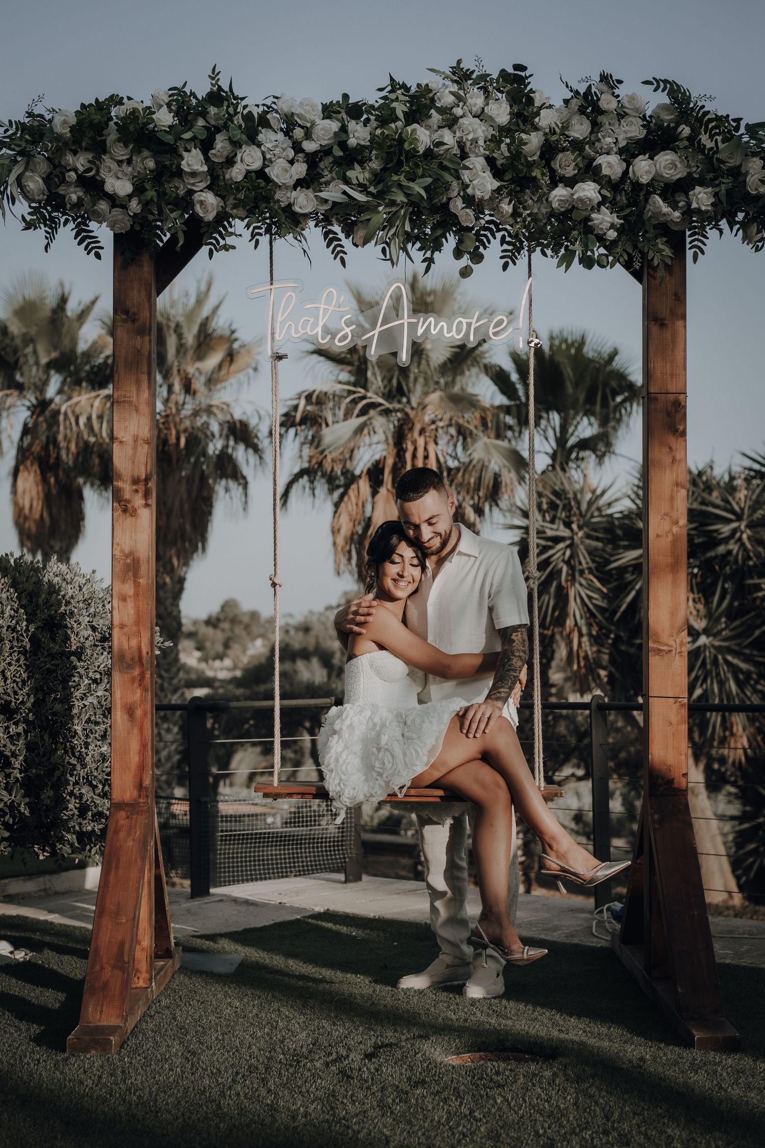 A romantic couple embraces on a swing beneath a white floral arch during sunset.