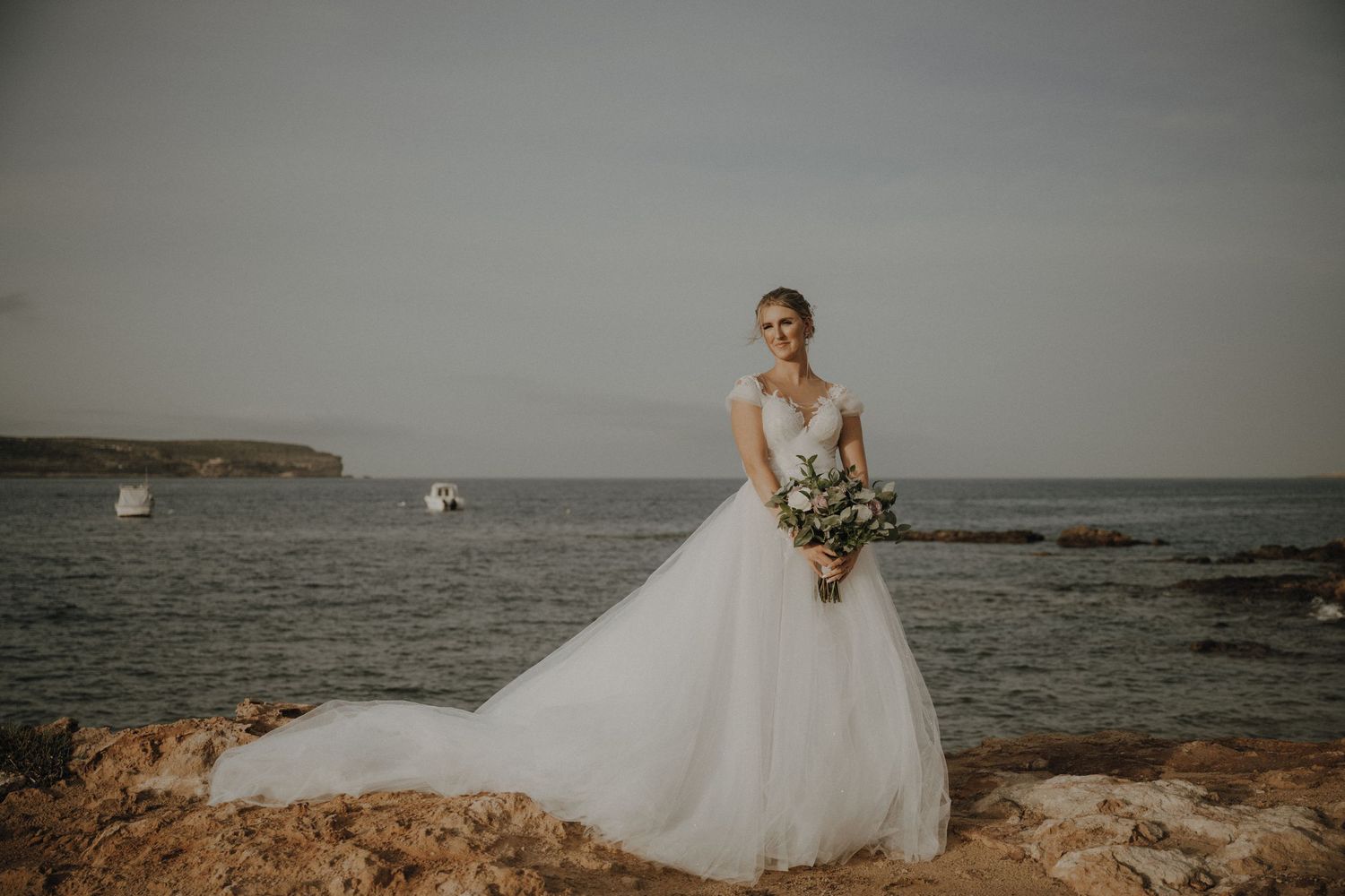 A bride in a flowing white wedding dress stands on a rocky coastal overlook with boats in the distance.