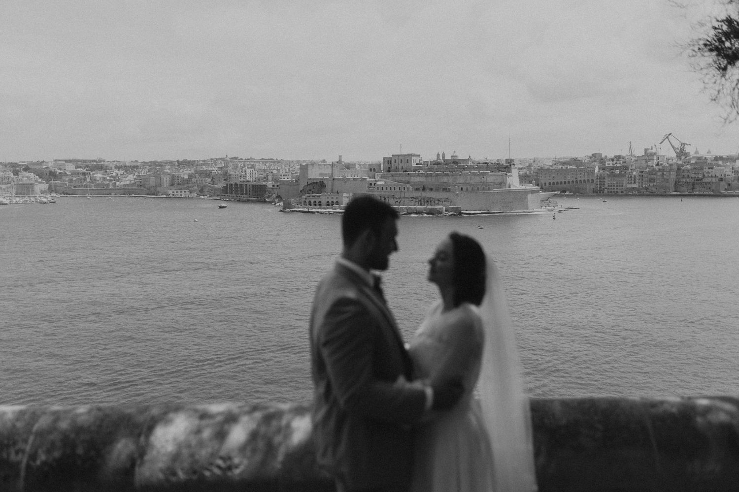 Wedding couple embracing by waterfront with cityscape in background.