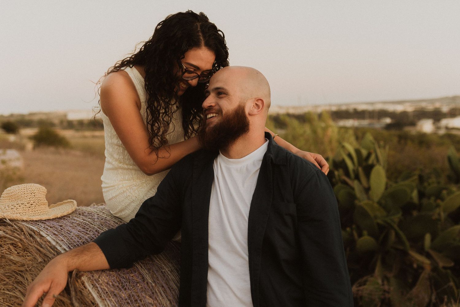 A romantic couple shares an intimate moment on a hilltop overlooking a distant town at sunset.