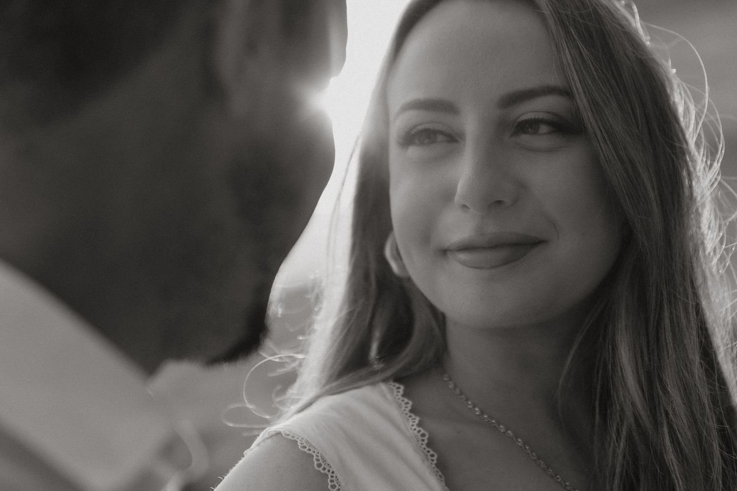 Black and white portrait of a smiling woman with long hair wearing a necklace, bathed in soft backlight.