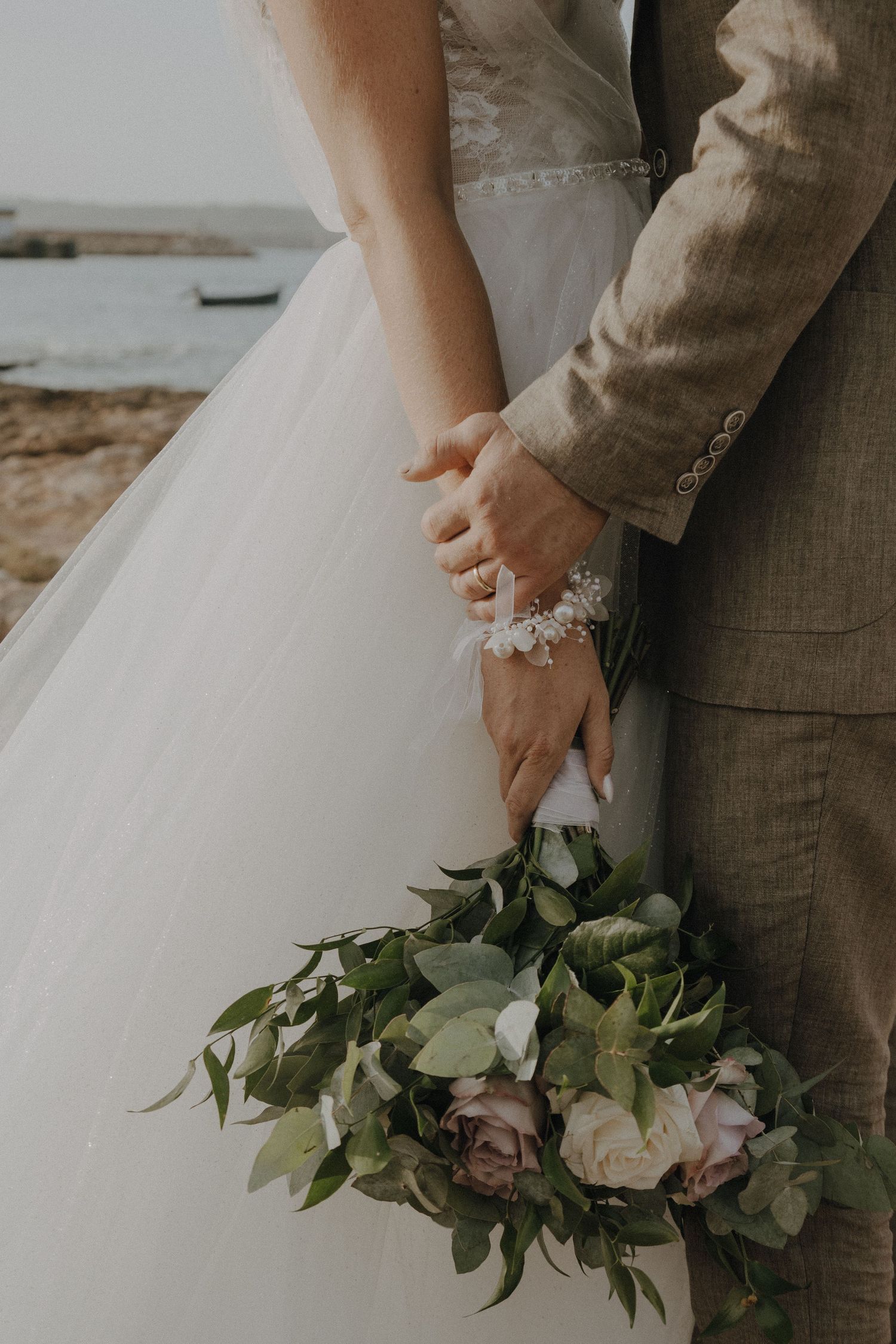 Close-up of newlyweds holding hands with a romantic bouquet of roses and greenery against a seaside backdrop.