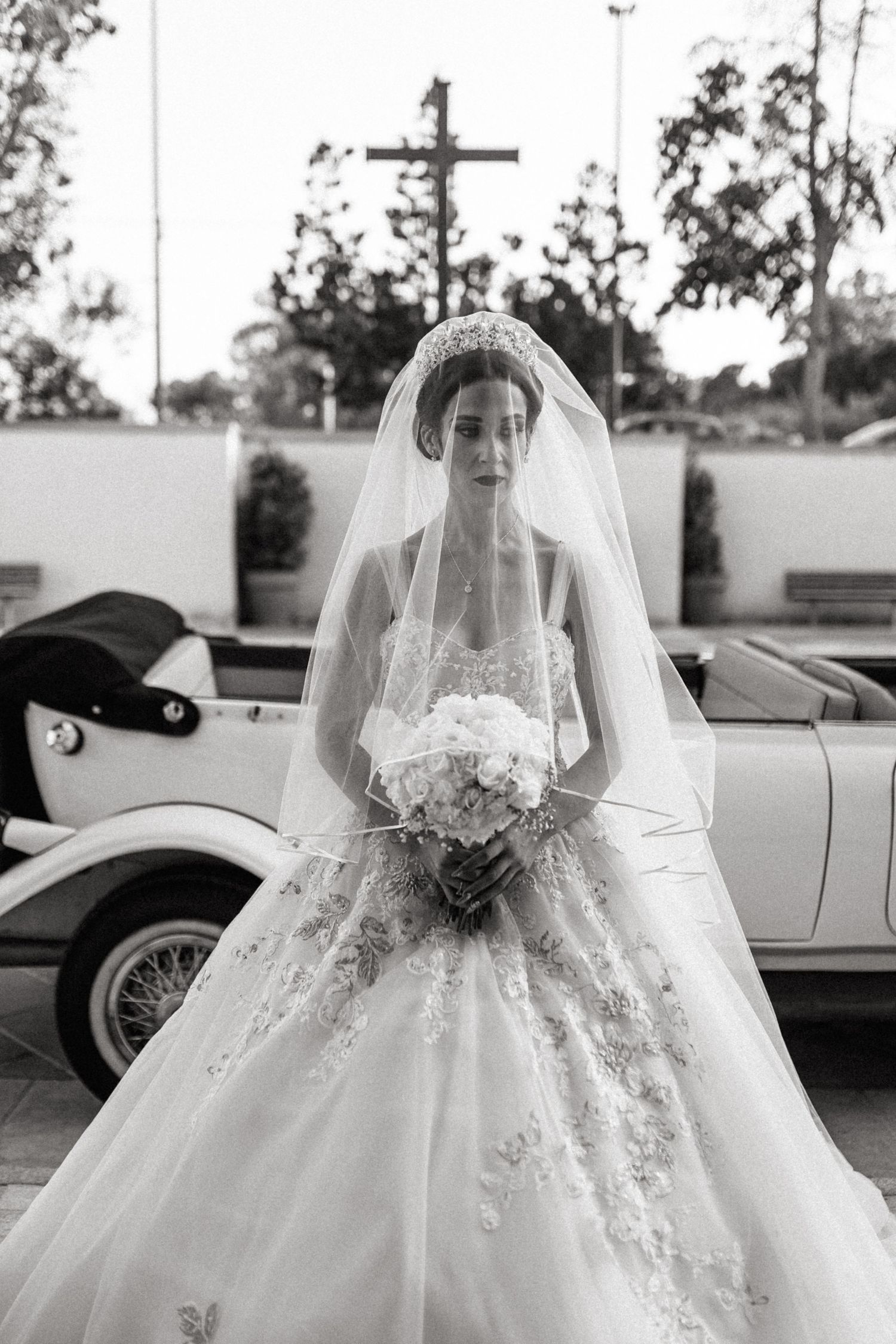 Elegant bride in lace ballgown poses with bouquet in front of church cross.