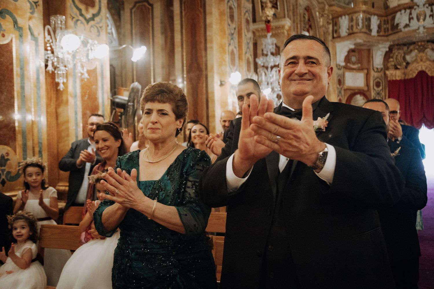 Wedding guests applaud during an elegant ceremony in an ornate church interior with chandeliers.