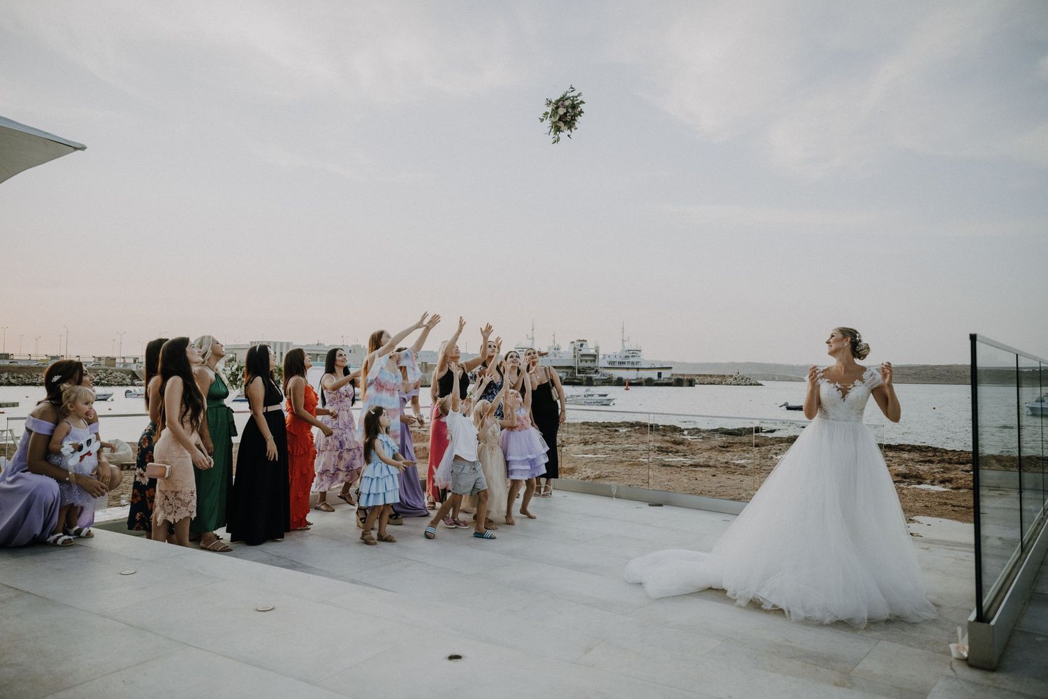 Guests reach for a flying bouquet during a waterfront wedding ceremony.