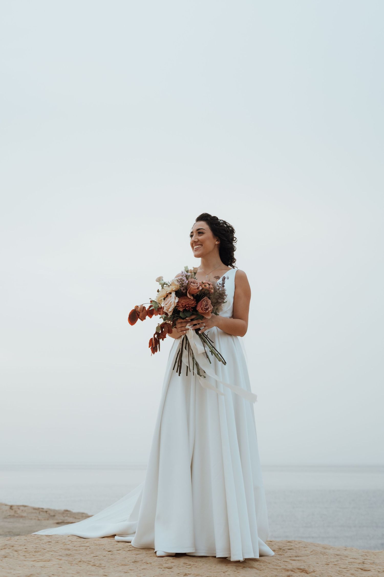 A bride in a flowing white gown holds a rich autumn-colored bouquet against a misty ocean backdrop.