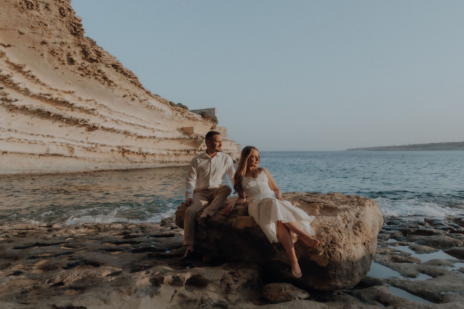 Couple sitting on a rock by the sea near white cliffs at dusk, dressed in elegant white outfits.