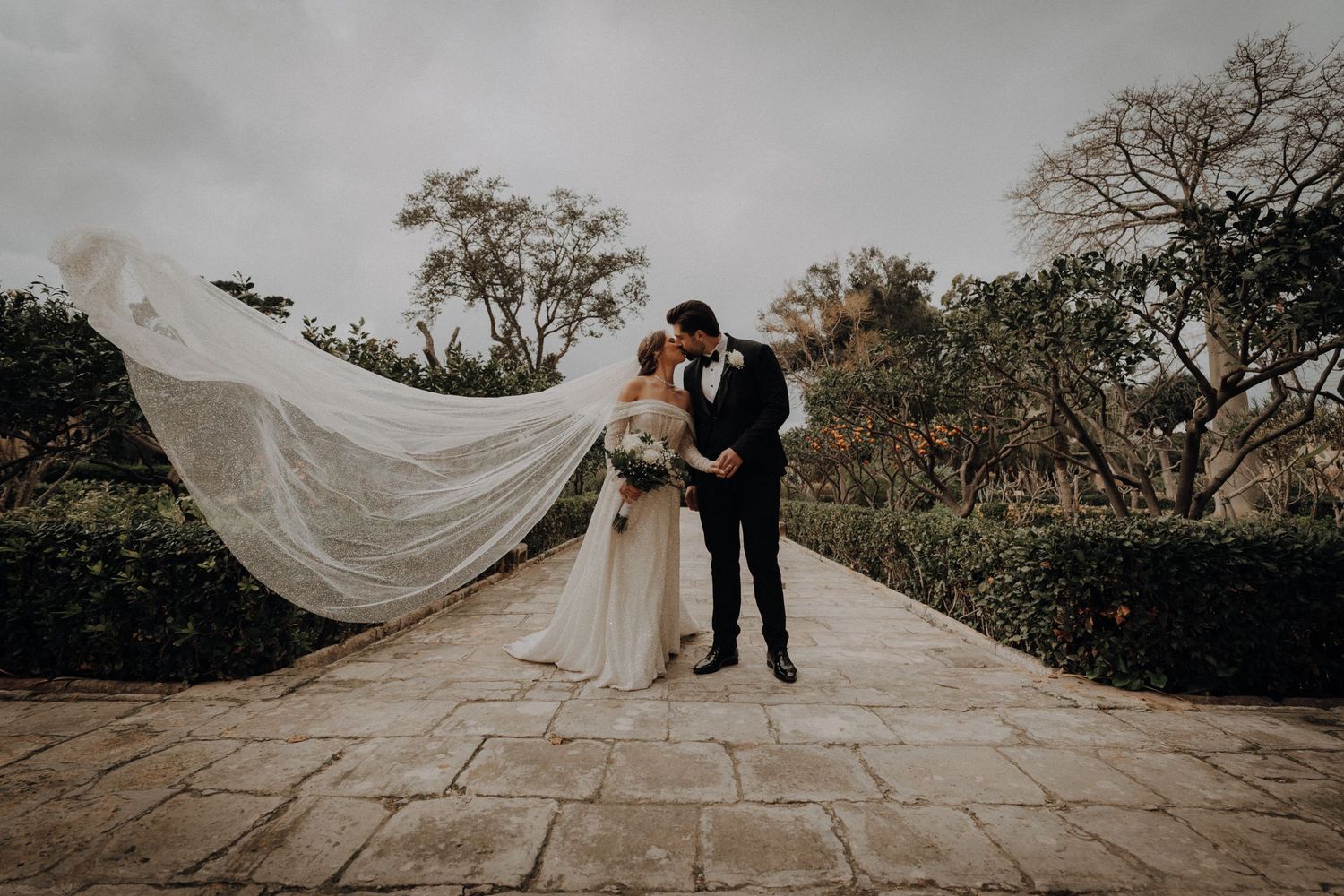 A flowing white wedding veil billows in the breeze on stone steps surrounded by greenery.