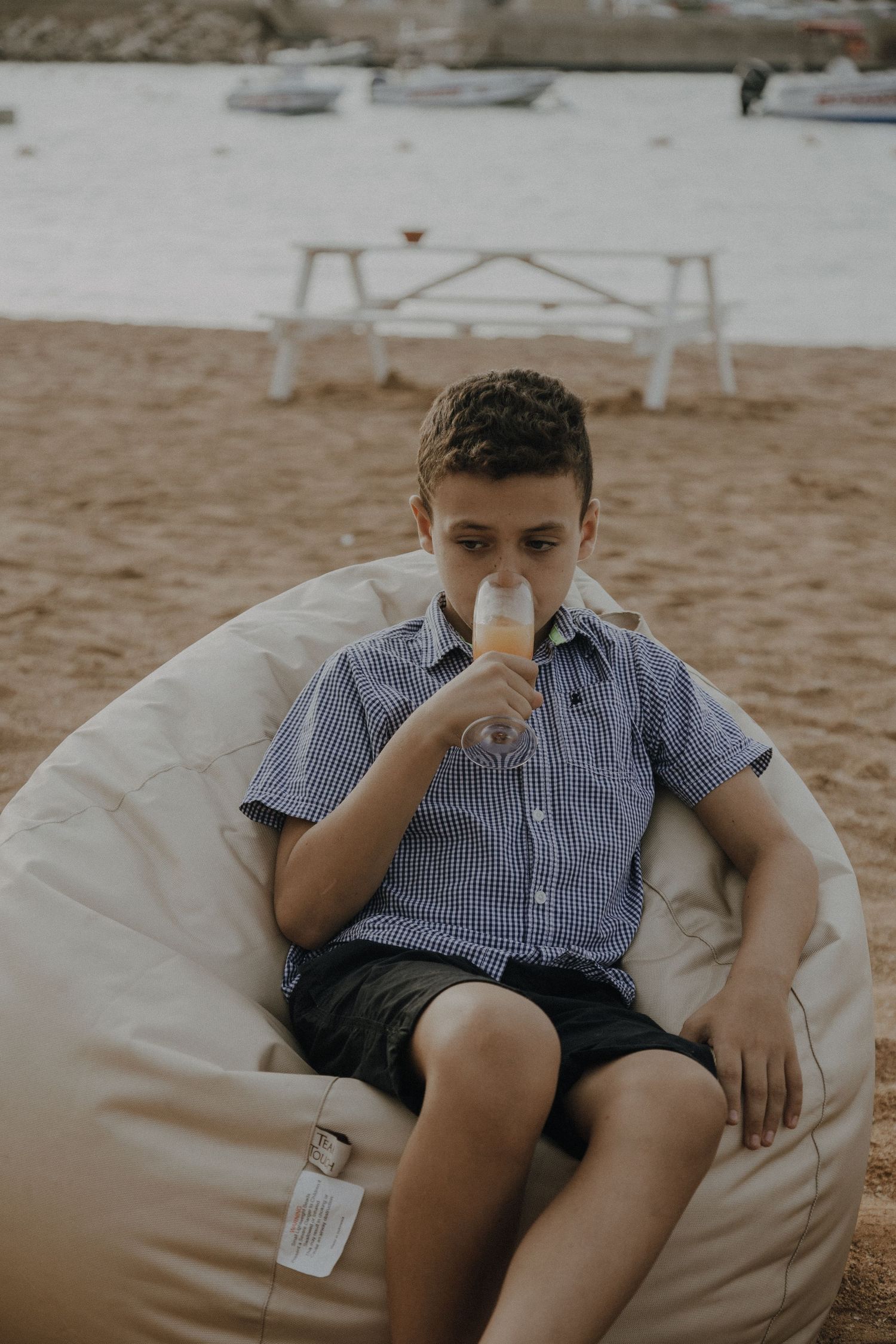 Young boy in checkered shirt sips drink while sitting in beige beanbag chair.