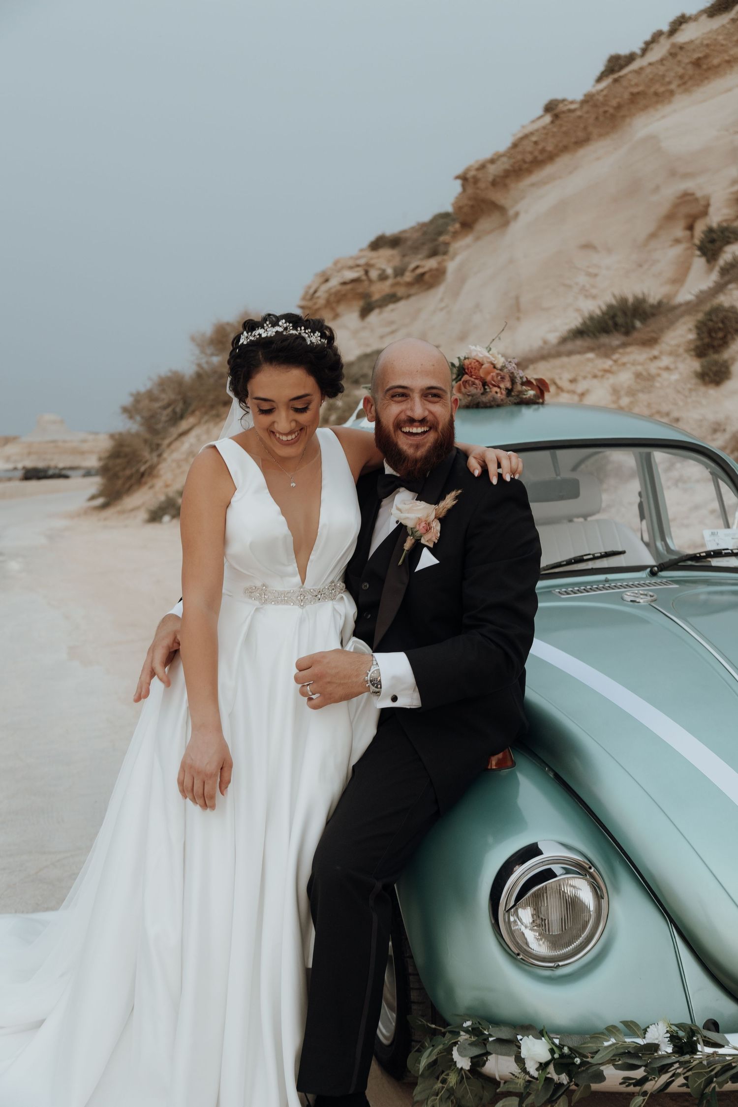 A happy couple poses by a vintage mint green car on their wedding day at a scenic coastal location.