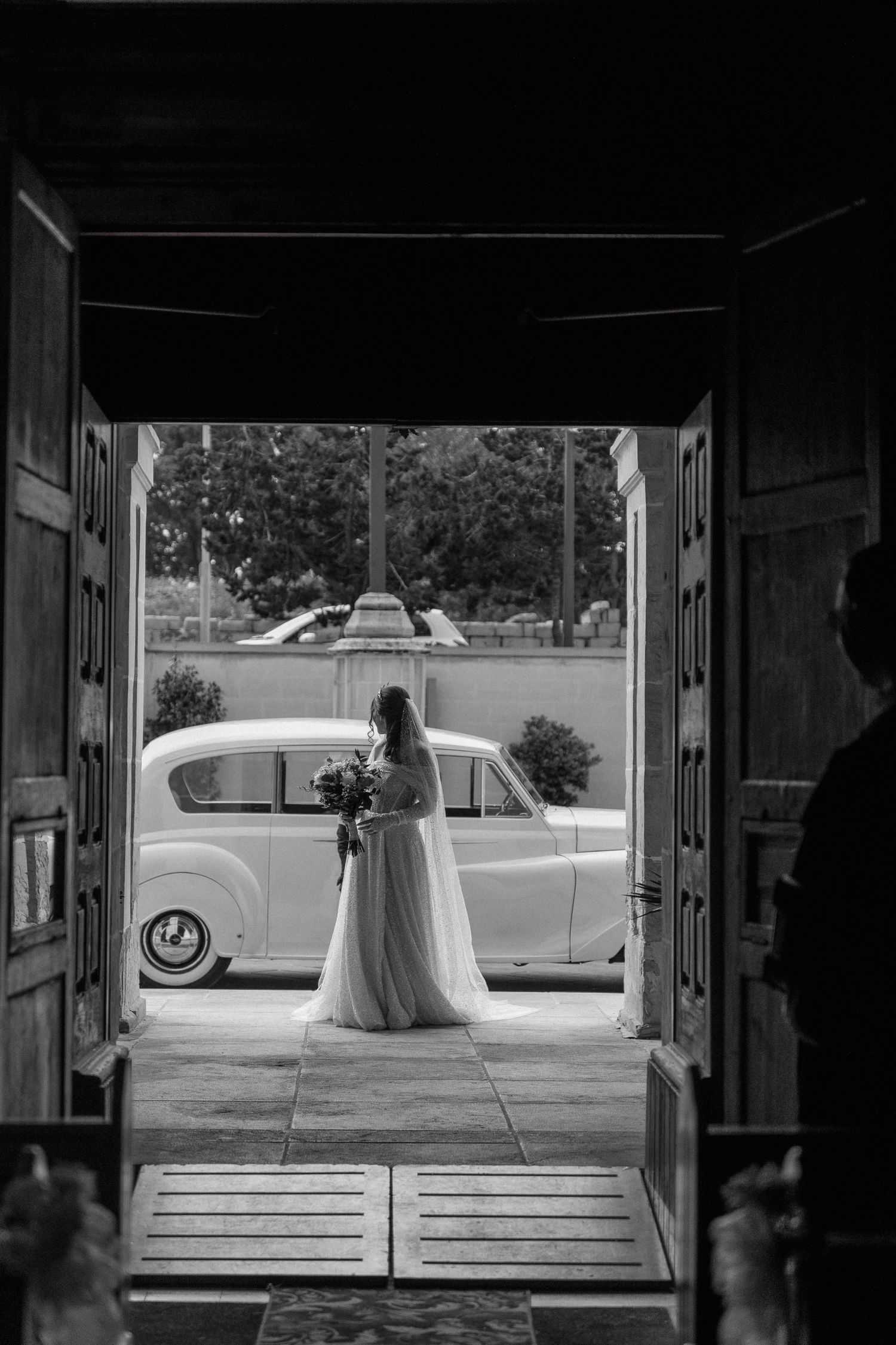 Black and white photo of bride silhouetted in doorway with vintage car in background.