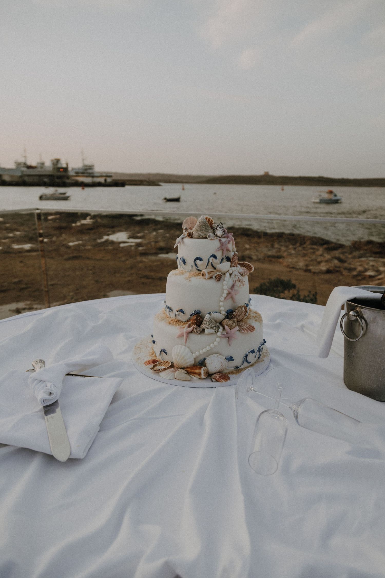 Wedding cake displayed on white tablecloth with ocean and boats in background.