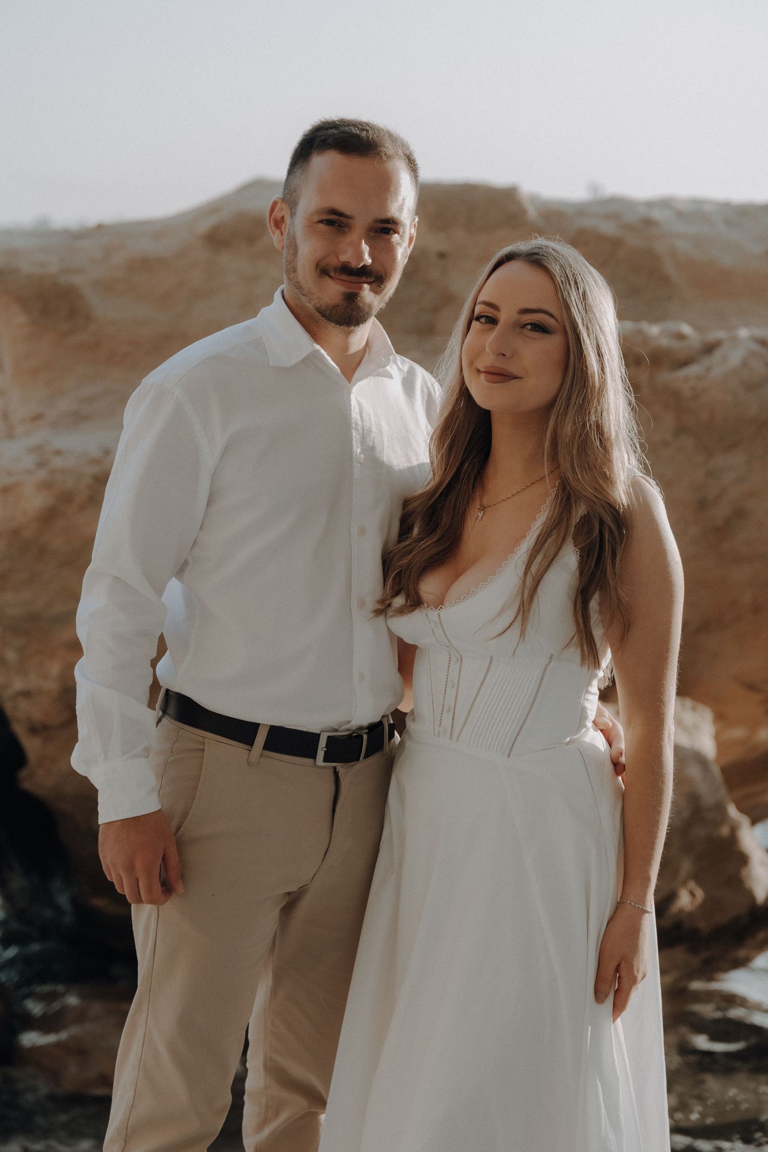 A couple in white attire poses romantically against rocky terrain, the woman in a white gown and man in a white shirt.