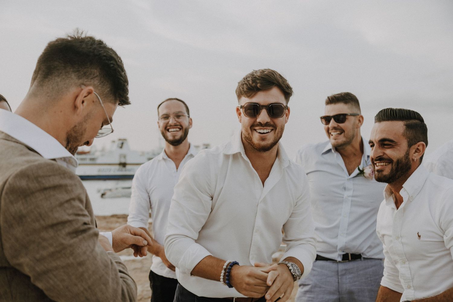 Groomsmen in white shirts and sunglasses share a laugh at seaside wedding.