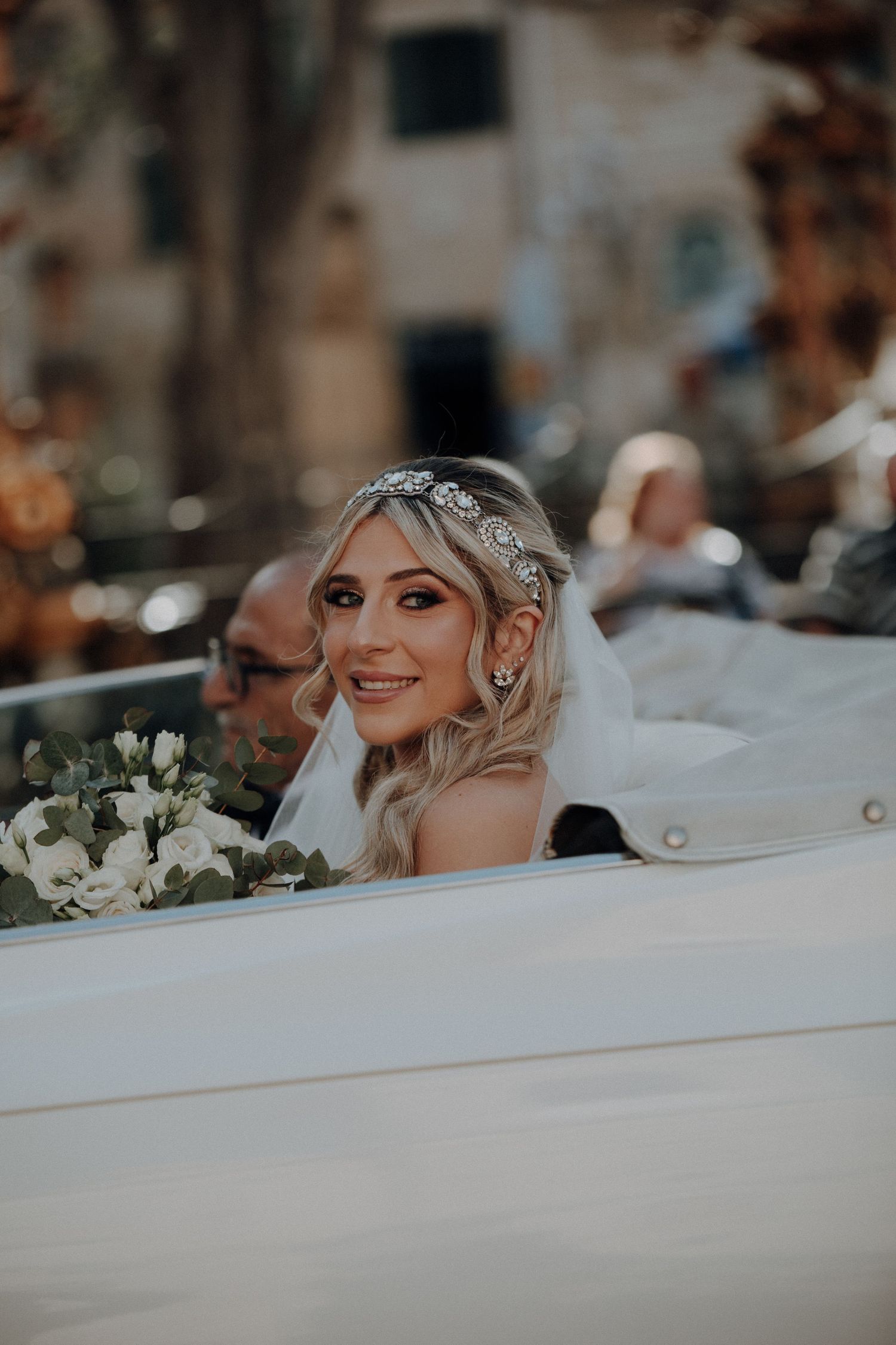A glamorous bride smiles from inside a white vintage car during her wedding celebration.