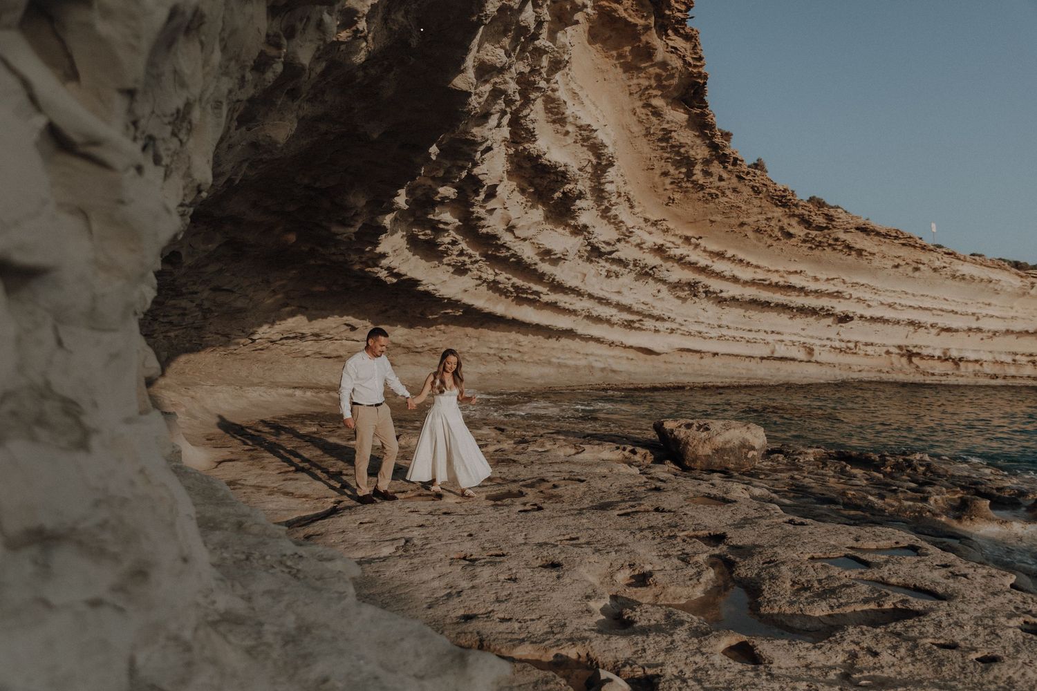 Couple in wedding attire walking along rocky coastal cliffs by the ocean during golden hour.