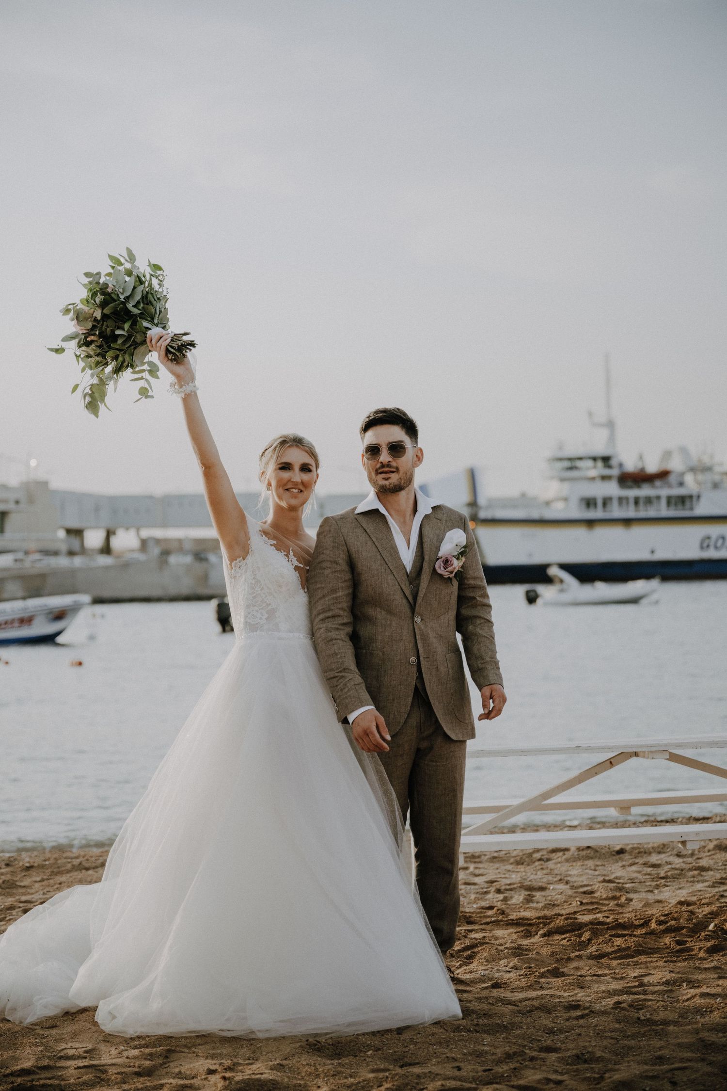 Newlyweds celebrate on beach with bride holding bouquet high and boats visible in harbor.