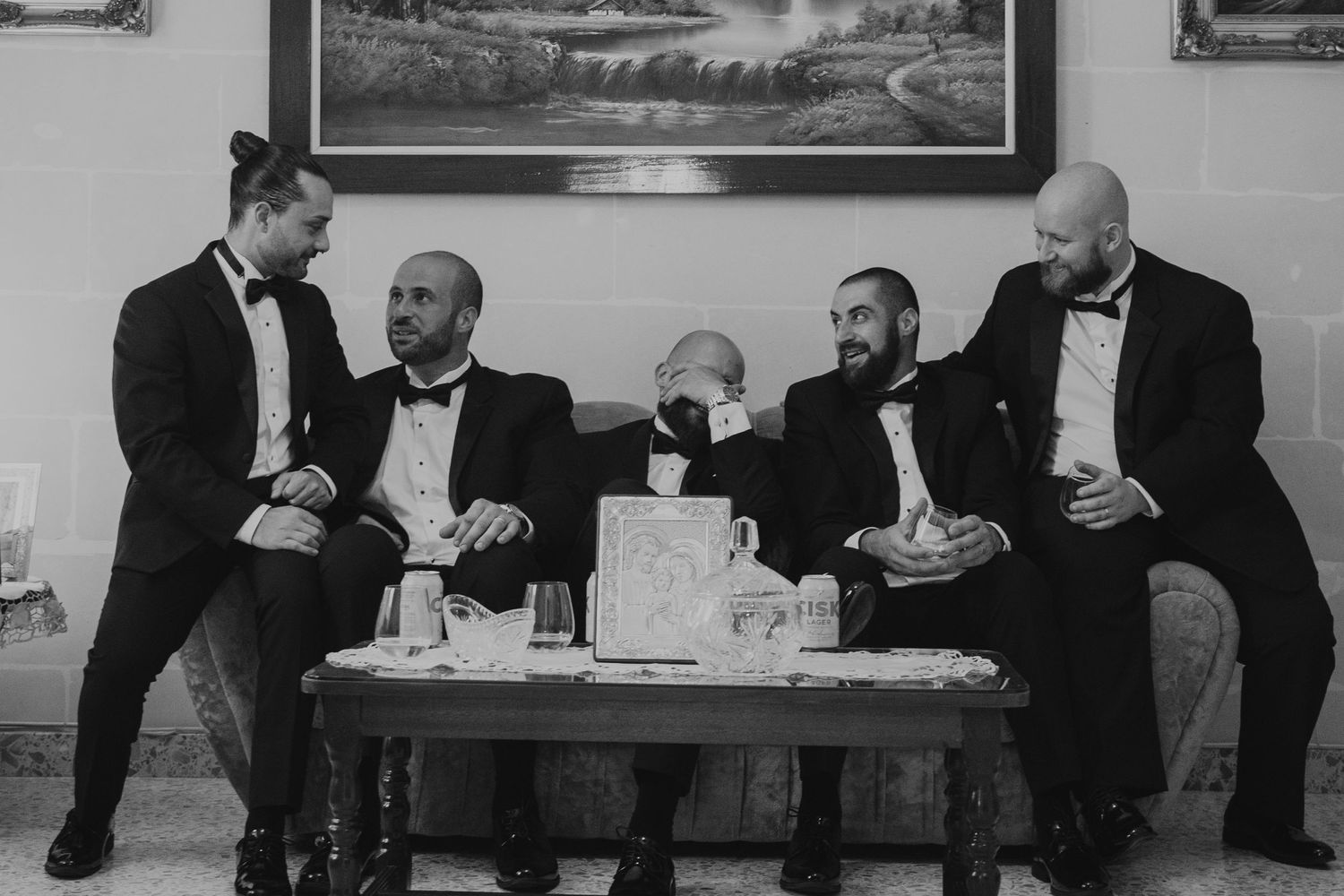 Men in tuxedos sitting and chatting on a bench against a wall with landscape paintings.