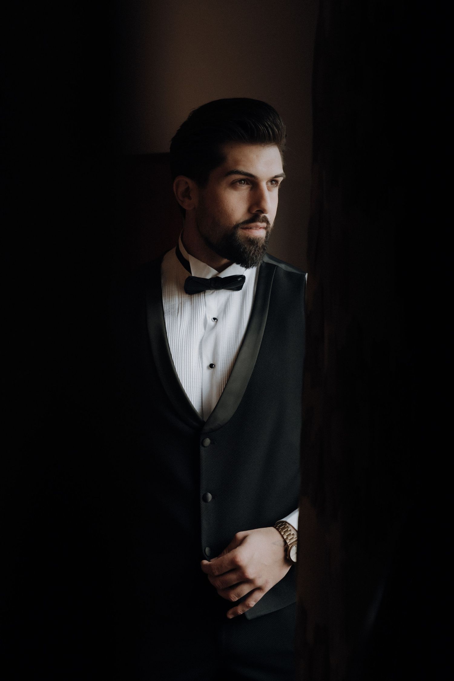 Dramatic portrait of groom in classic black tuxedo and white dress shirt against dark background.