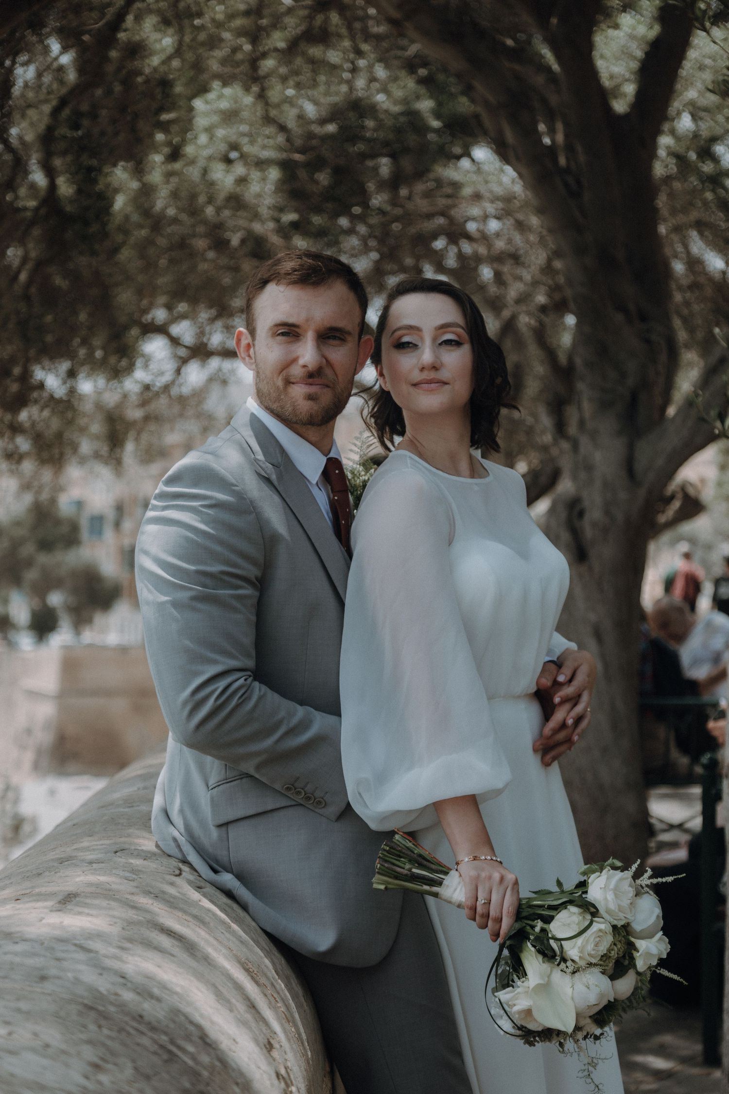 Elegant couple in wedding attire posing outdoors with white bouquet.
