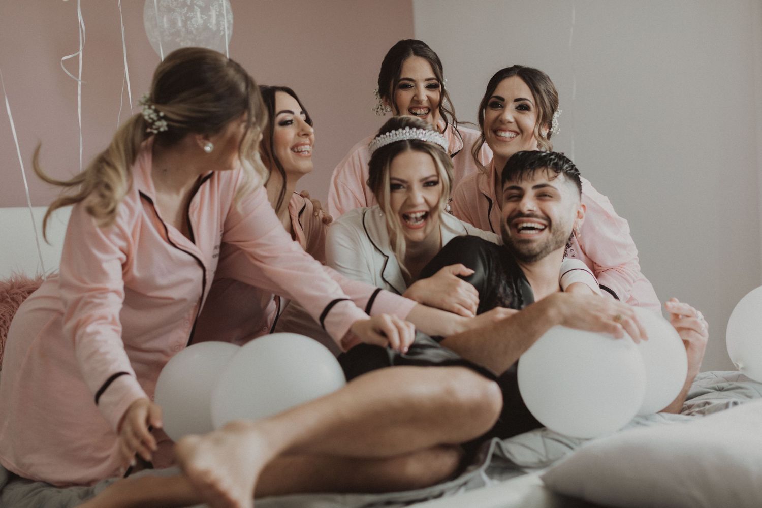 Playful wedding party scene with bridesmaids in pink loungewear relaxing on bed with balloons.