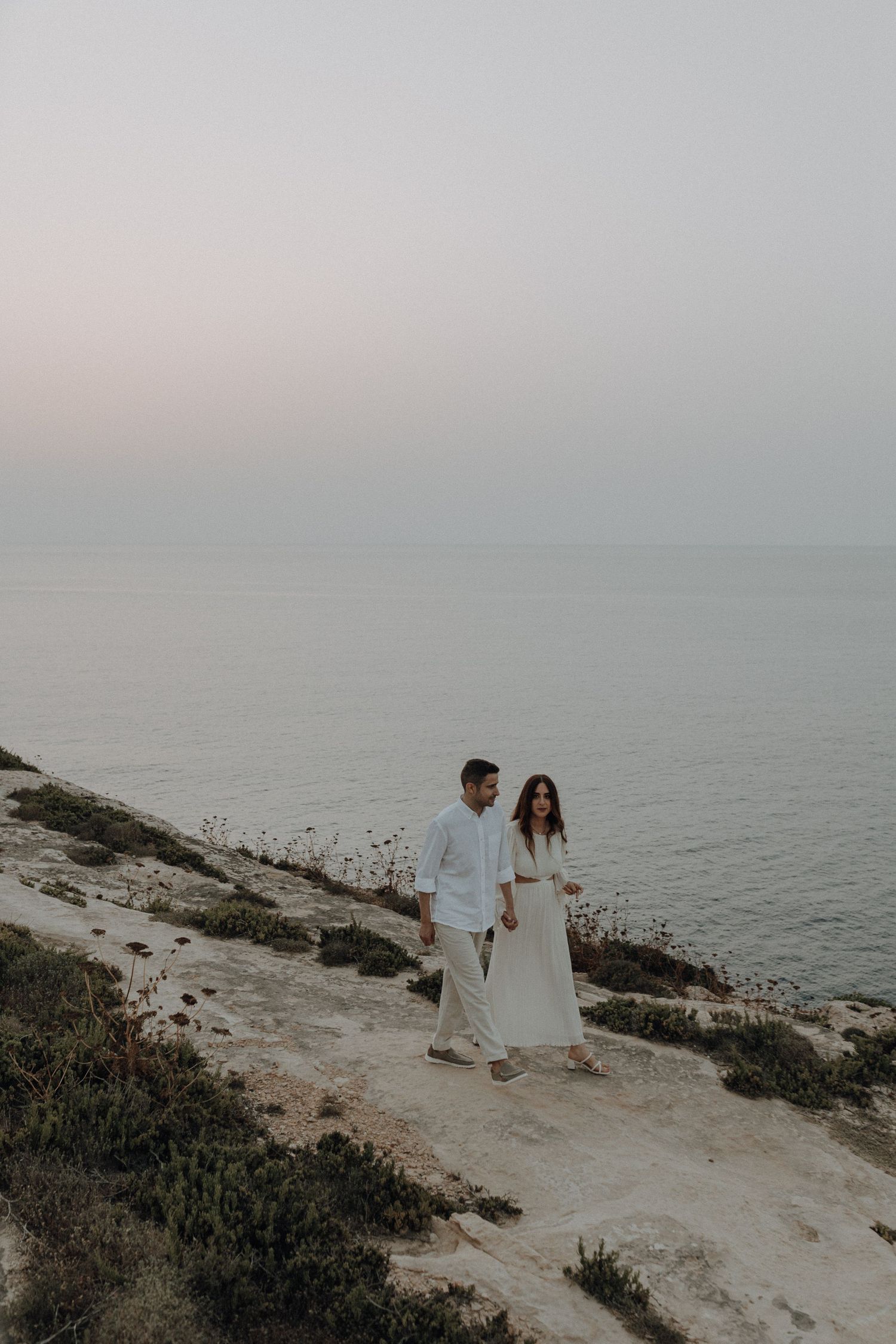 A couple in white clothing walks along a coastal path overlooking the ocean.