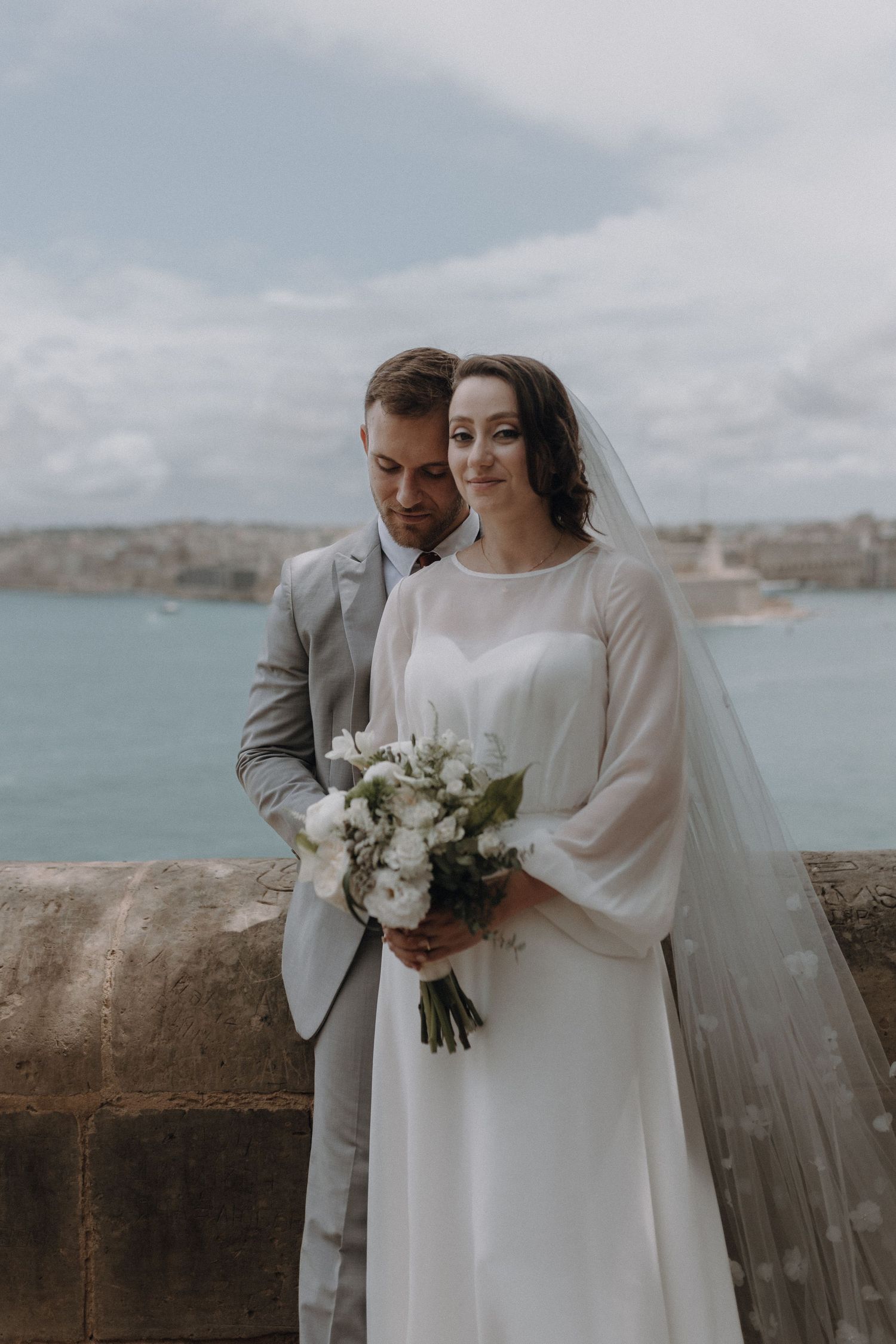 A wedding couple poses against a coastal backdrop with white bouquet and elegant attire.