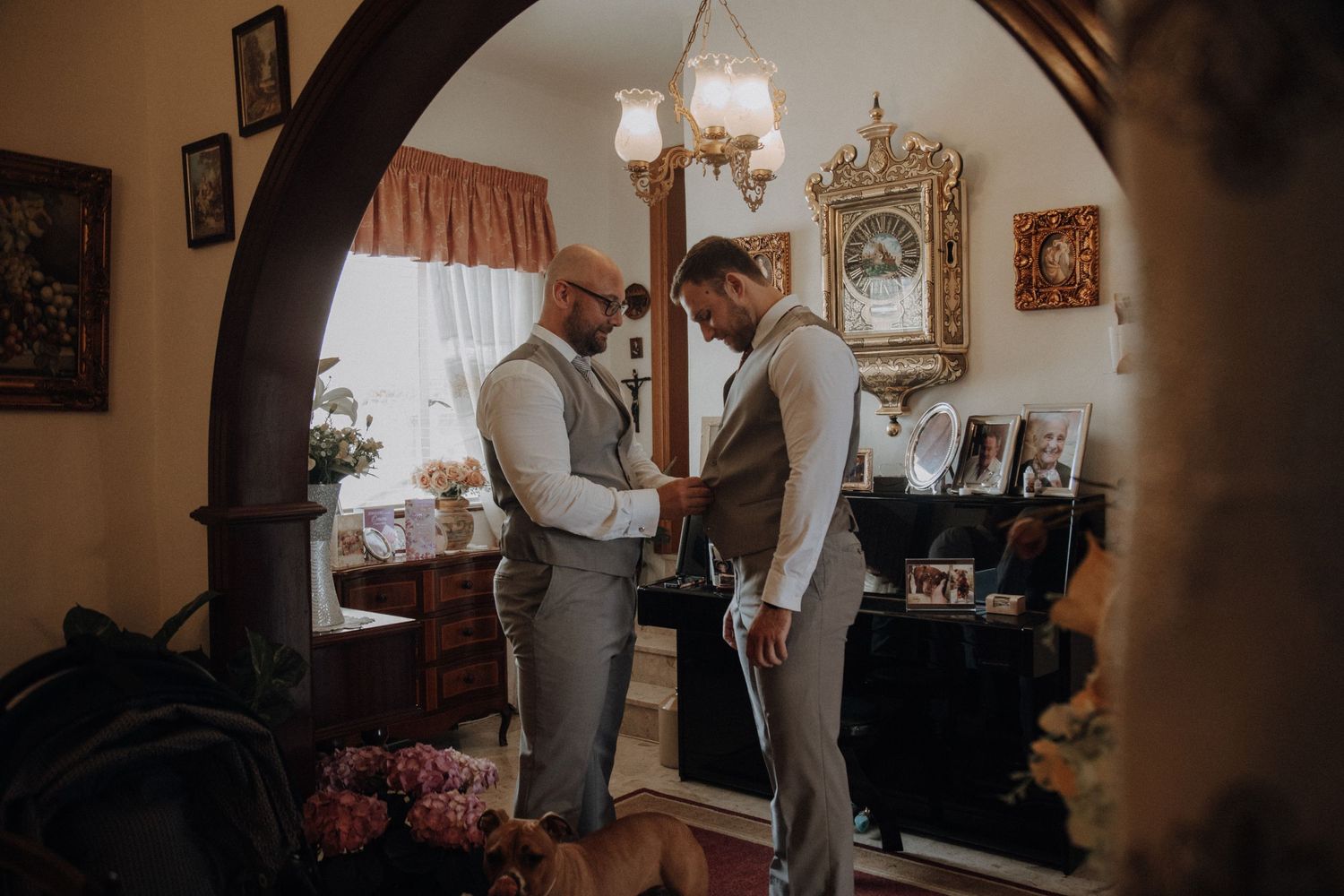 Two people in grey suits help each other with formal attire under an ornate archway.
